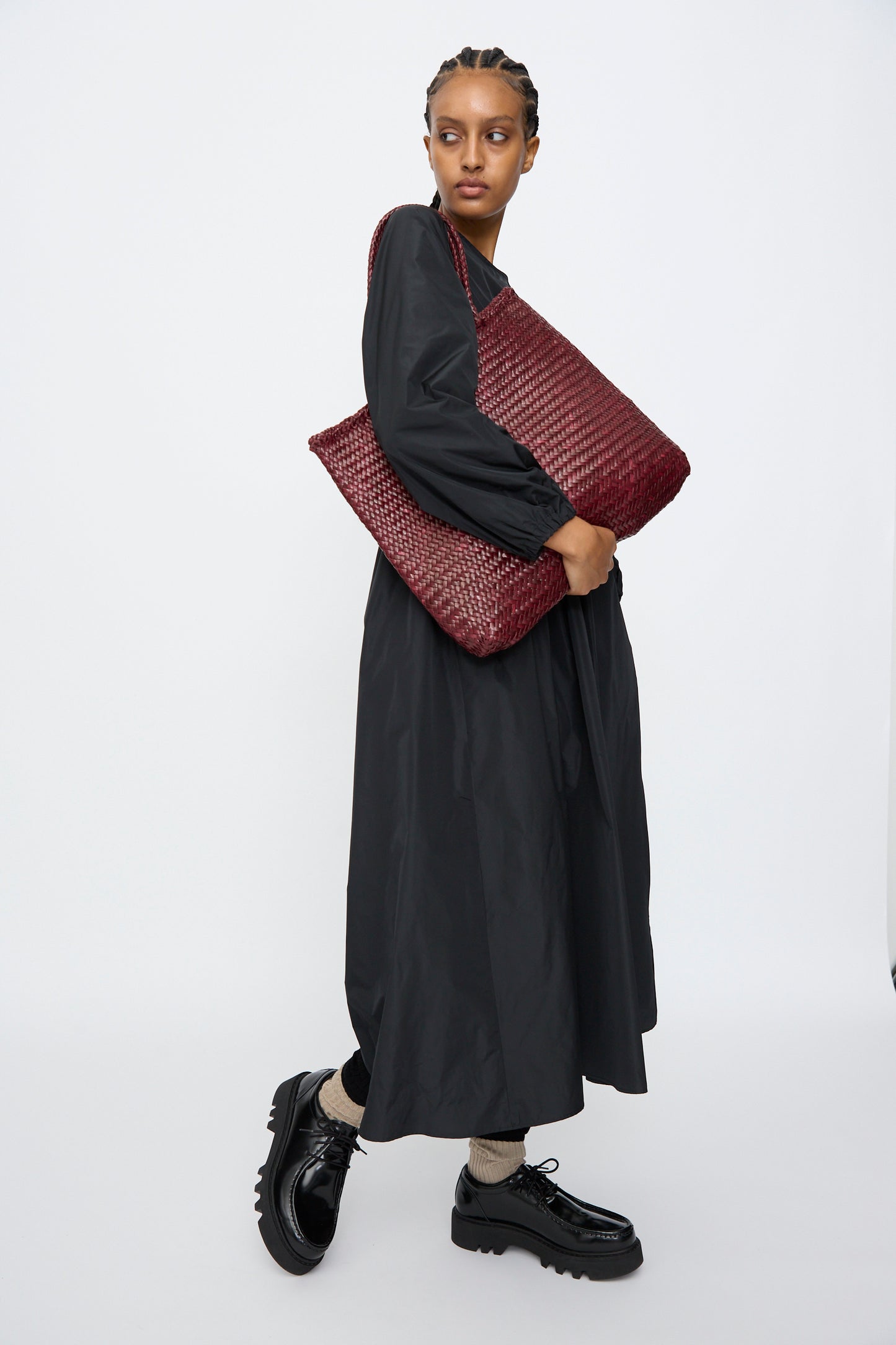 A person in a long black dress and black platform shoes holds the Dragon Diffusion Sophie Large in Bordo tote, made from buffalo leather, against a plain white background.