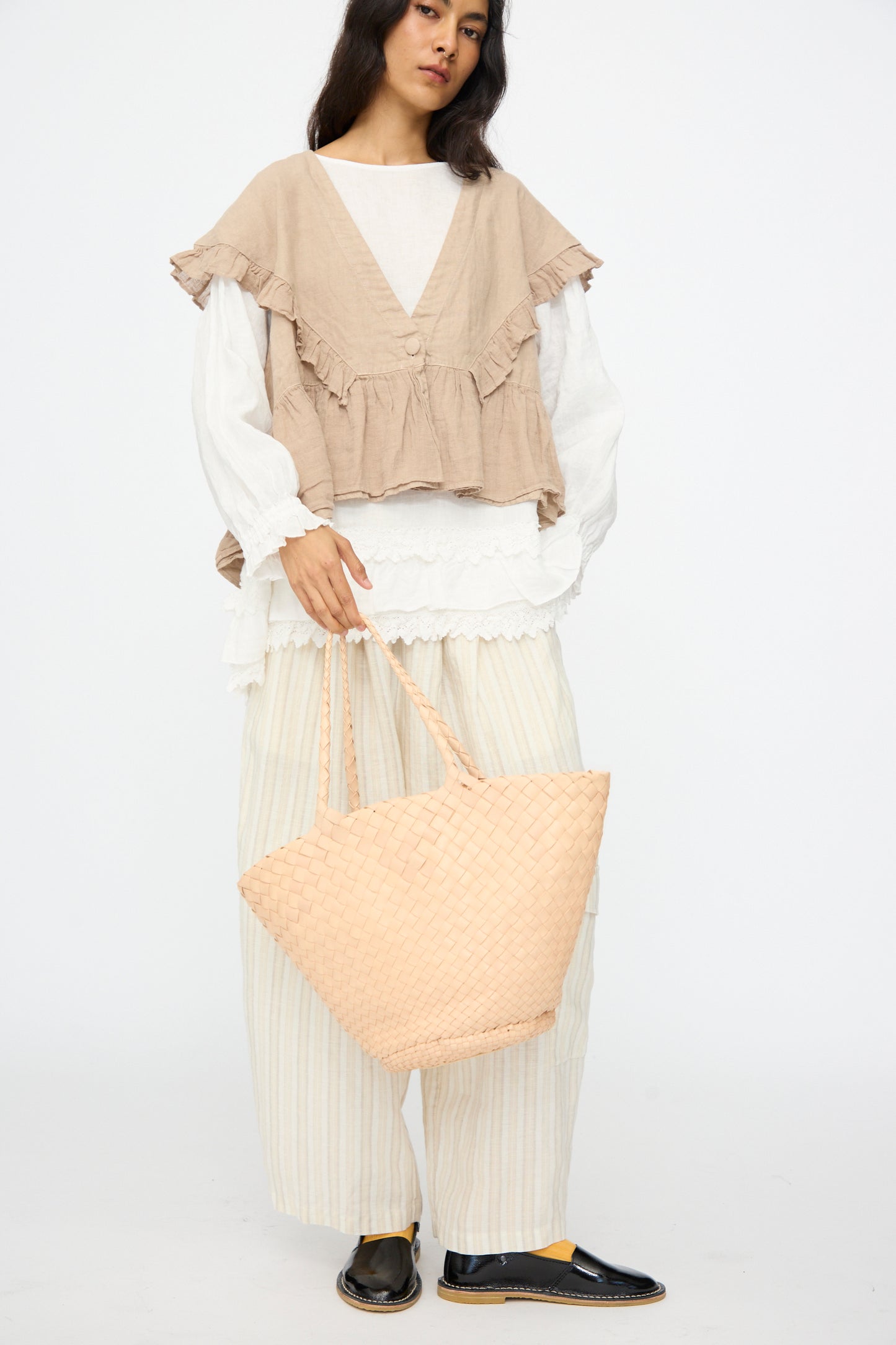 A person stands against a plain white background, holding the Dragon Diffusion Egola in Natural woven leather handbag, styled with a beige ruffled vest, white blouse, wide striped pants, and black shoes.