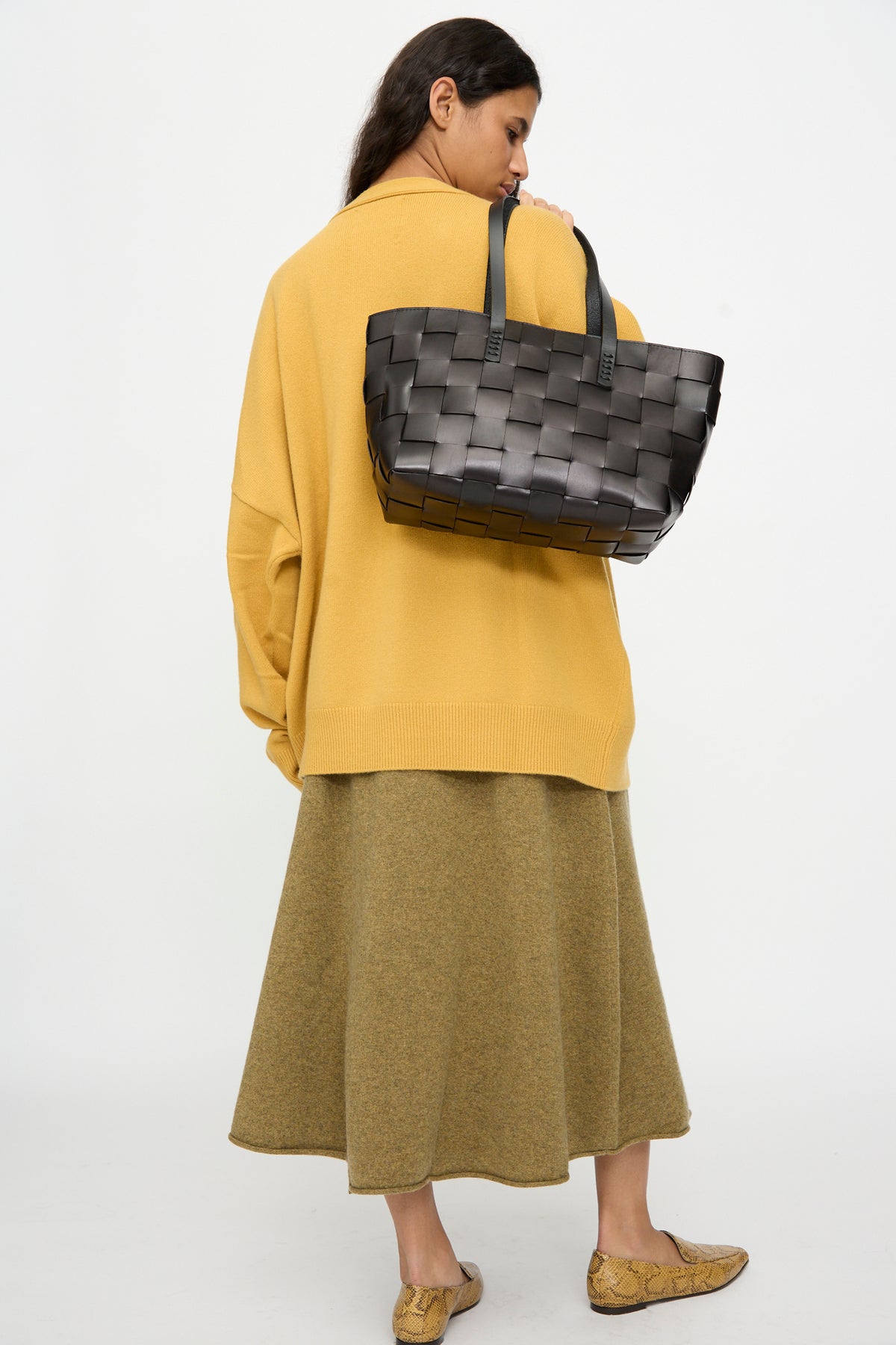 A woman in a yellow sweater and olive skirt stands with her back to the camera, carrying the Dragon Diffusion Japan Tote in Black—a cross-woven leather handbag in a basket shape—on her shoulder.