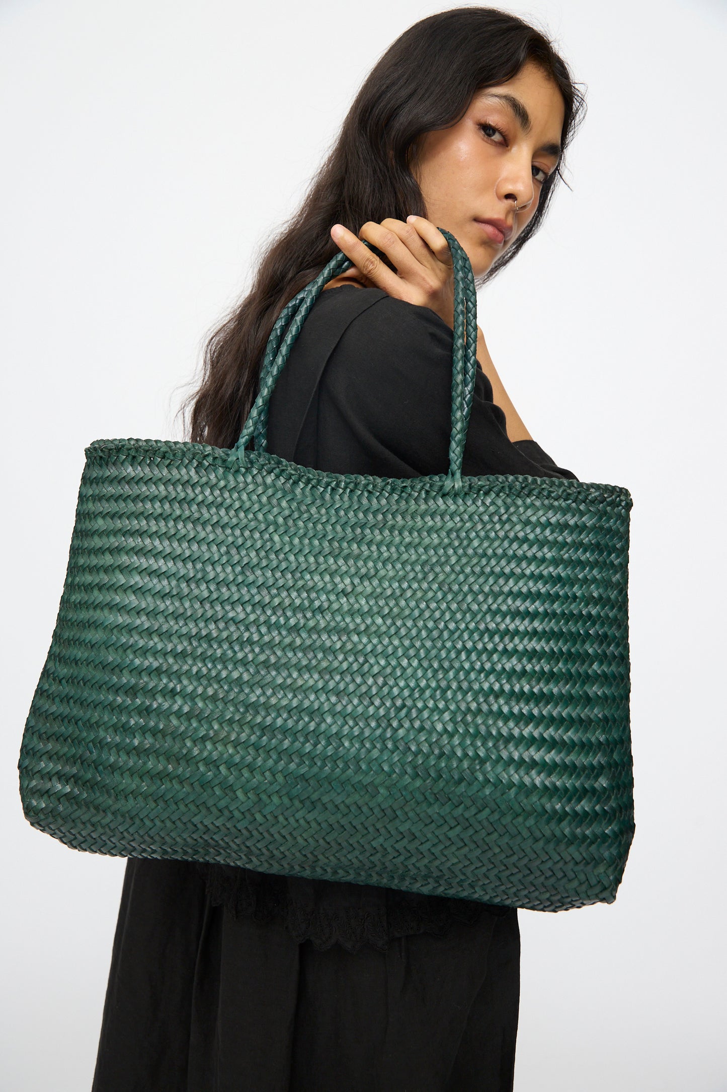 A woman in black holds the Dragon Diffusion Sophie Large in Forest, a dark green handwoven tote with braided handles, over her shoulder against a plain white background.