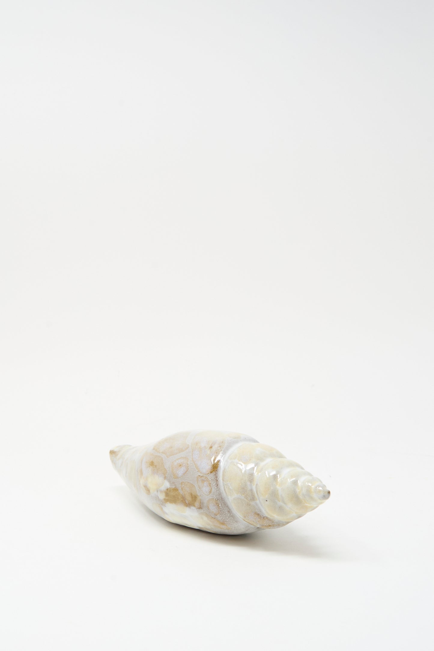 The E.C.C.E Shell Rattle XCVII, a handmade conical ceramic shell with a spiral tip and light brown spots, is displayed on a plain white background.