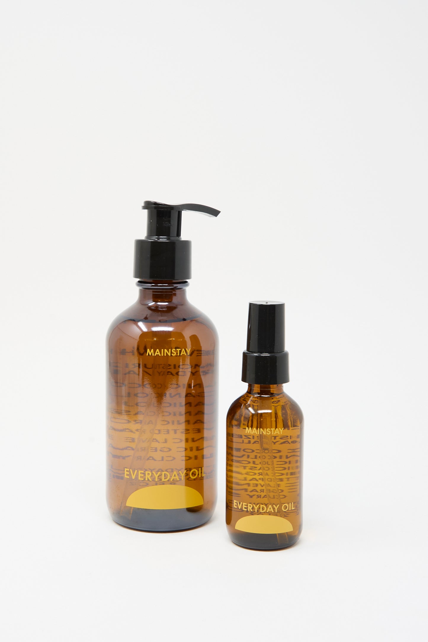 Two amber bottles of Everyday Oil's Mainstay Blend (8 oz)—one with a pump and one with a spray top—are displayed against a white background, highlighting this all-natural skincare essential.