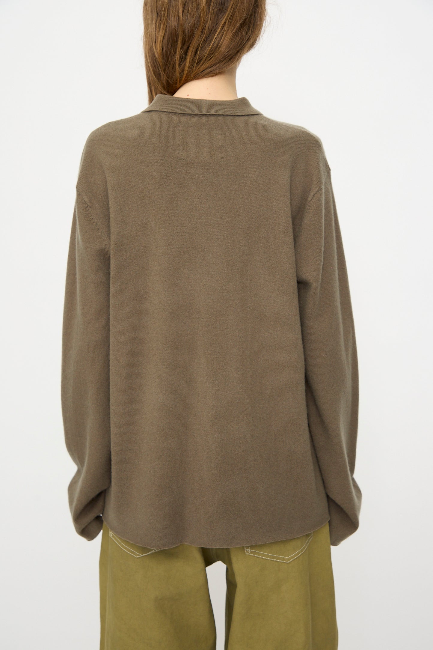 A person with long brown hair is shown from the back, wearing the Extreme Cashmere No. 199 Alligator Sweater in Army and olive green pants, set against a plain white background.