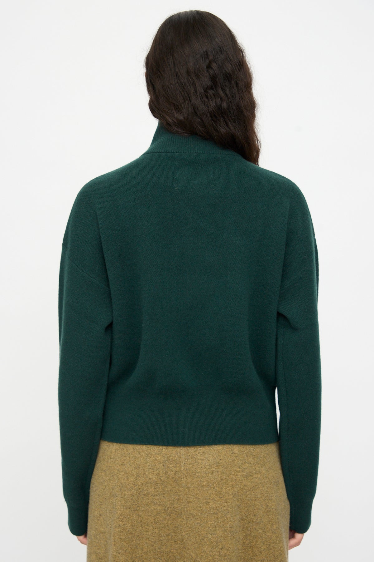 A person with long, wavy brown hair is shown from the back against a plain white background, wearing the Extreme Cashmere No. 385 Xtra Cute Cardigan in Forest paired with a light brown skirt.