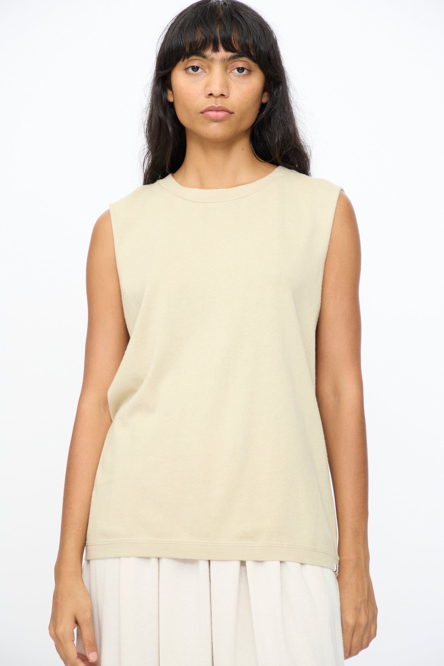 A woman with long dark hair wears the Extreme Cashmere No. 408 Muscle Tee in Desert and a light skirt, standing against a plain white background.