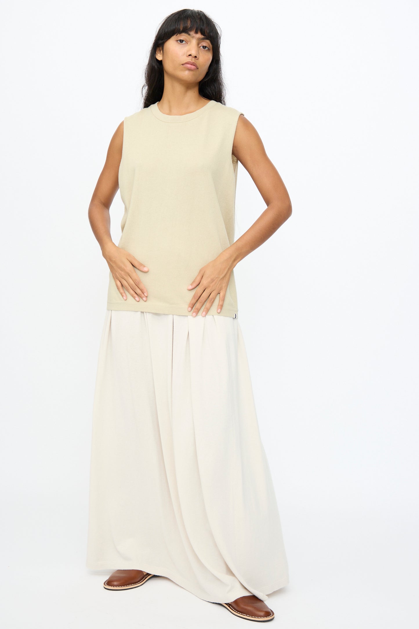 A woman stands against a plain white background wearing Extreme Cashmere's No. 408 Muscle Tee in Desert, paired with a long cream skirt and brown shoes.