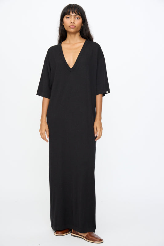 A person stands against a plain background wearing Extreme Cashmere's No. 410 Inda Dress in Raven—an ankle-length, relaxed-fit black t-shirt dress with short sleeves and a deep V-neck—paired with brown shoes.
