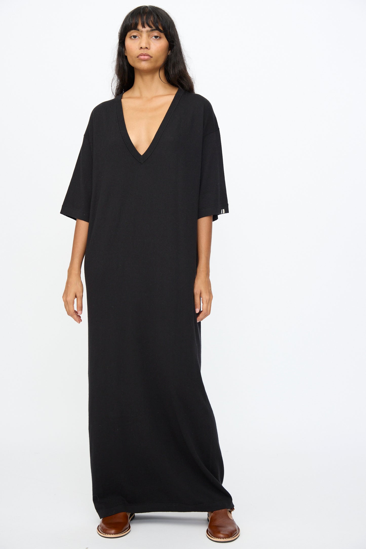 A person stands against a plain background wearing the Extreme Cashmere No. 410 Inda Dress in Raven, featuring a relaxed silhouette, V-neck, short sleeves, and paired with brown slip-on shoes.