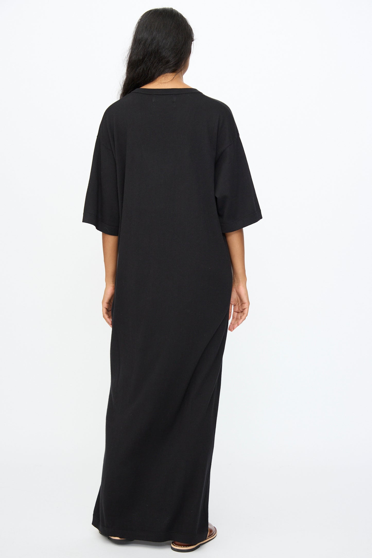 A person faces away from the camera in Extreme Cashmere's No. 410 Inda Dress in Raven—a black, relaxed-fit maxi t-shirt dress—paired with sandals, set against a plain white background.