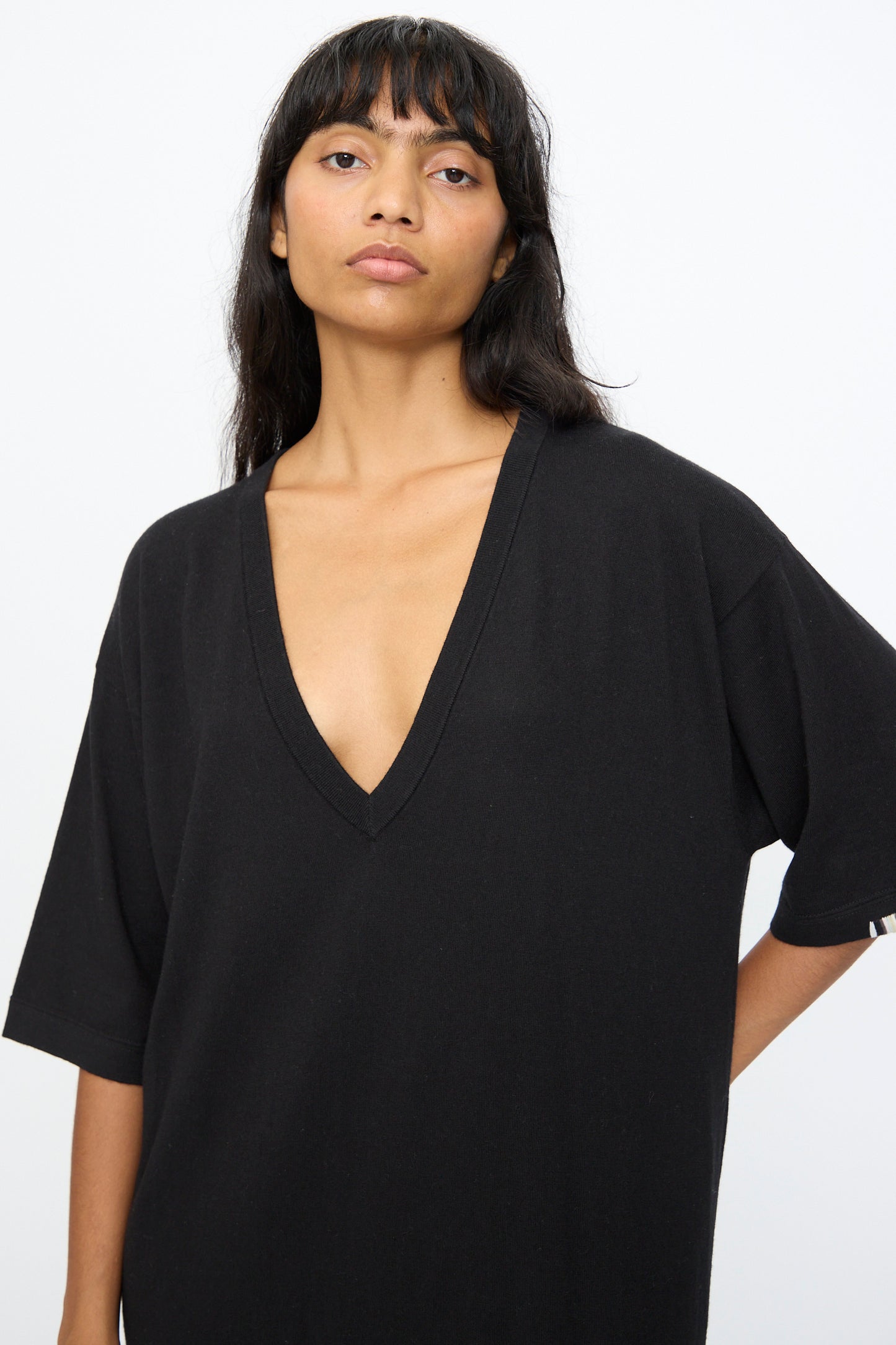 A woman with long dark hair wears the Extreme Cashmere No. 410 Inda Dress in Raven—a loose-fitting black V-neck maxi t-shirt dress with a relaxed silhouette—against a plain white background.