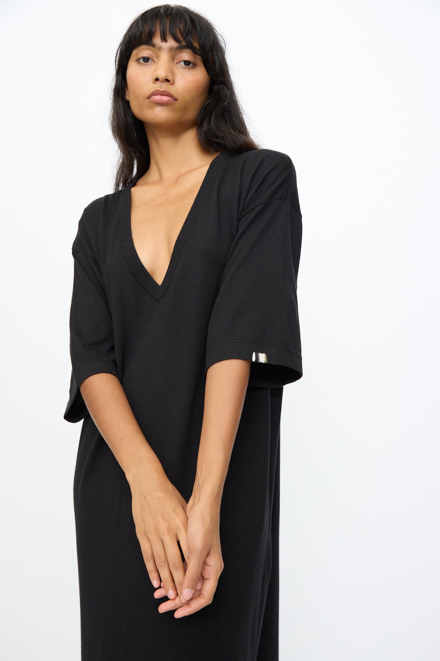 A person with long dark hair wears the No. 410 Inda Dress in Raven by Extreme Cashmere, a relaxed silhouette maxi t-shirt dress with a deep V-neckline and elbow-length sleeves, standing against a plain white background.