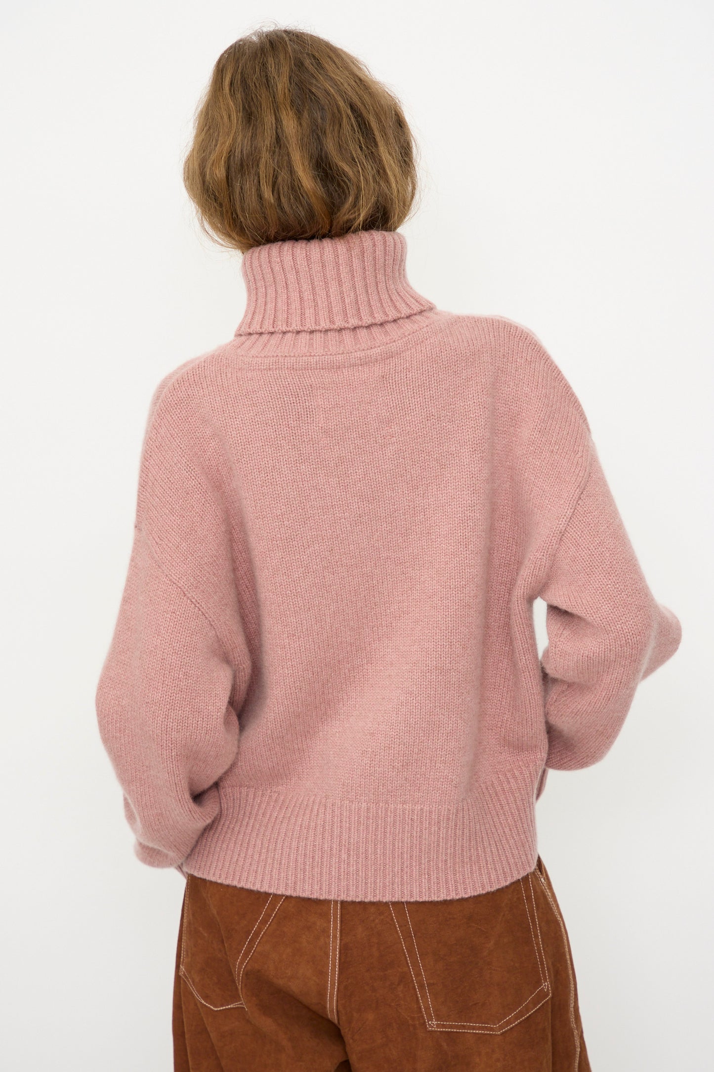A person with short, wavy light brown hair models the Extreme Cashmere No. 434 Crop Xtra Sweater in Moor with brown cropped pants, facing away from the camera against a plain white background.