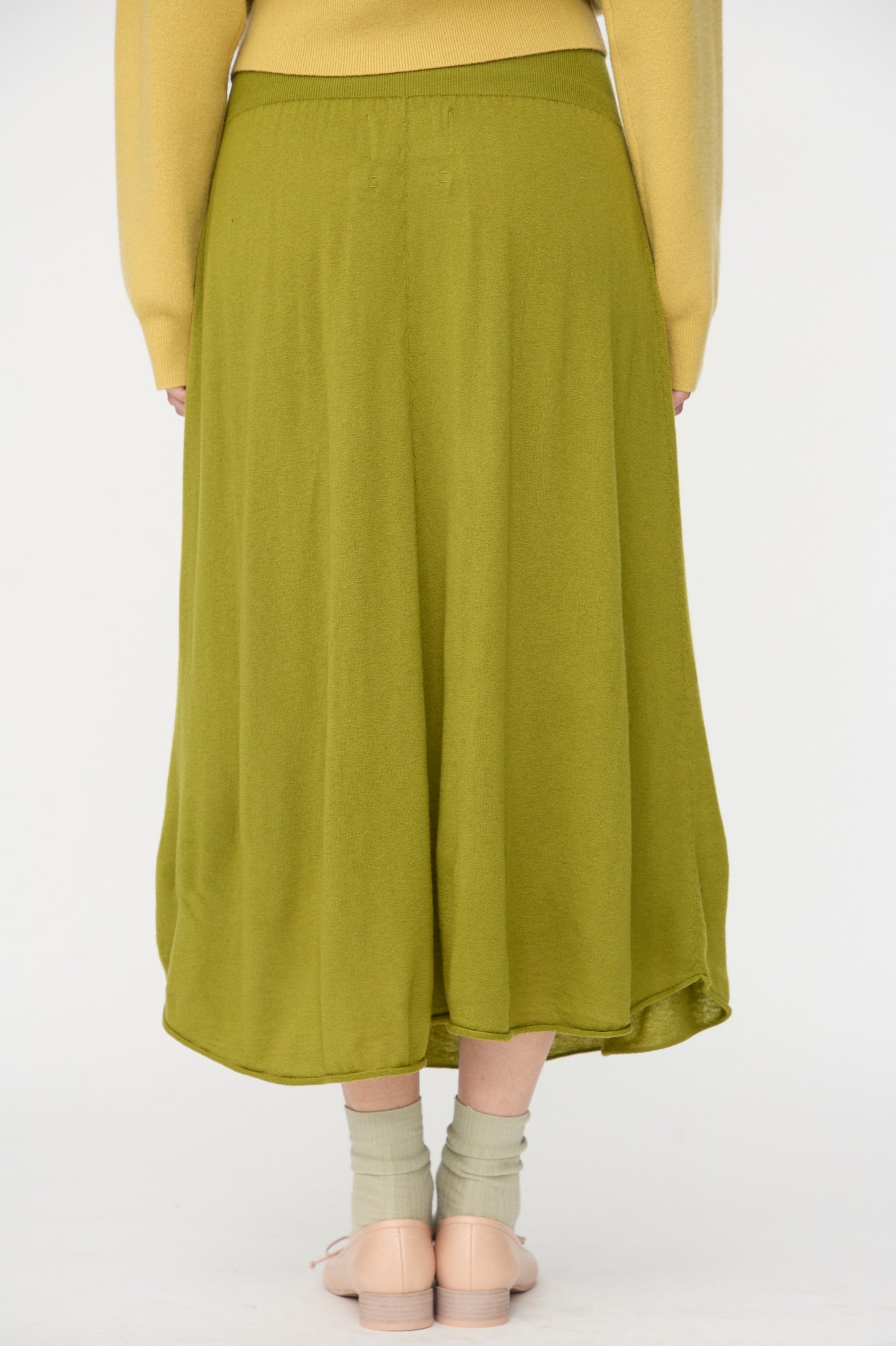 Model wearing the No. 470 Seduce Skirt in Grass by Extreme Cashmere with light green socks, beige shoes, and a yellow long-sleeve top, shown from the back on a plain white background.
