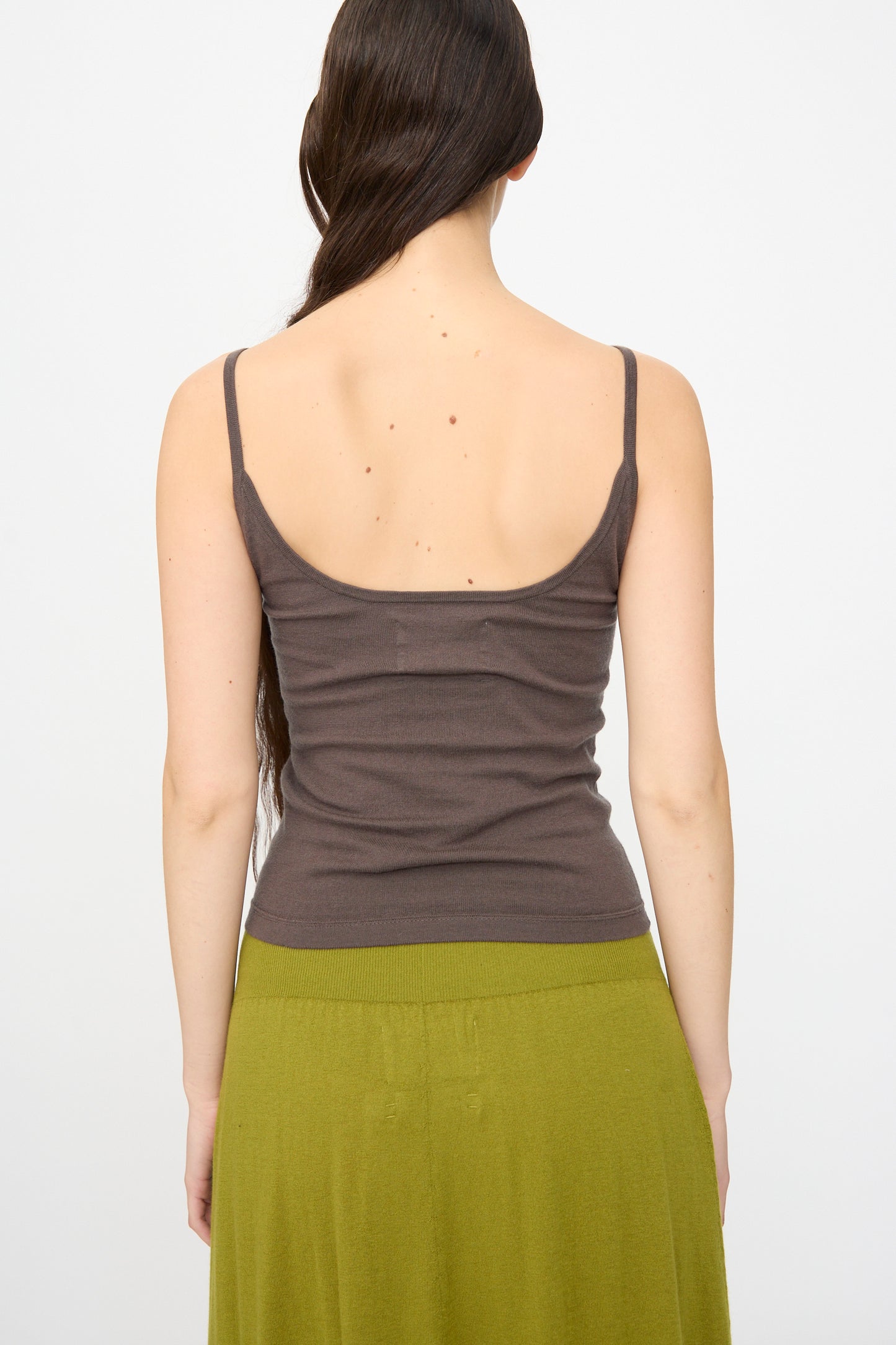 A person with long brown hair is seen from behind wearing the Extreme Cashmere No. 471 Britney Tank in Mud and a green skirt against a plain background.