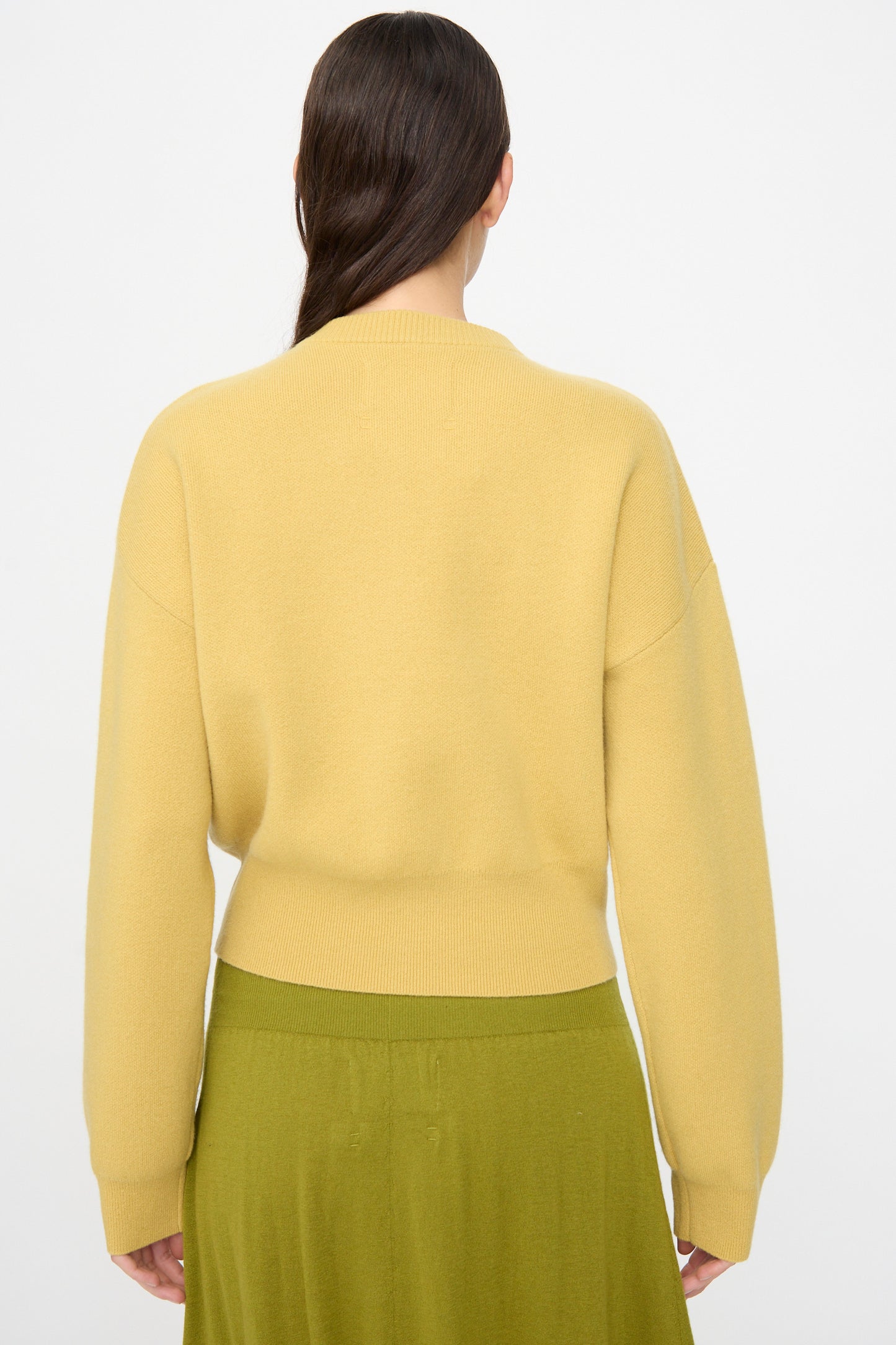 Person with long brown hair wears the Extreme Cashmere No. 478 Xtra Fun Cardigan in Corn and an olive green skirt, standing with back to the camera against a plain, light background.