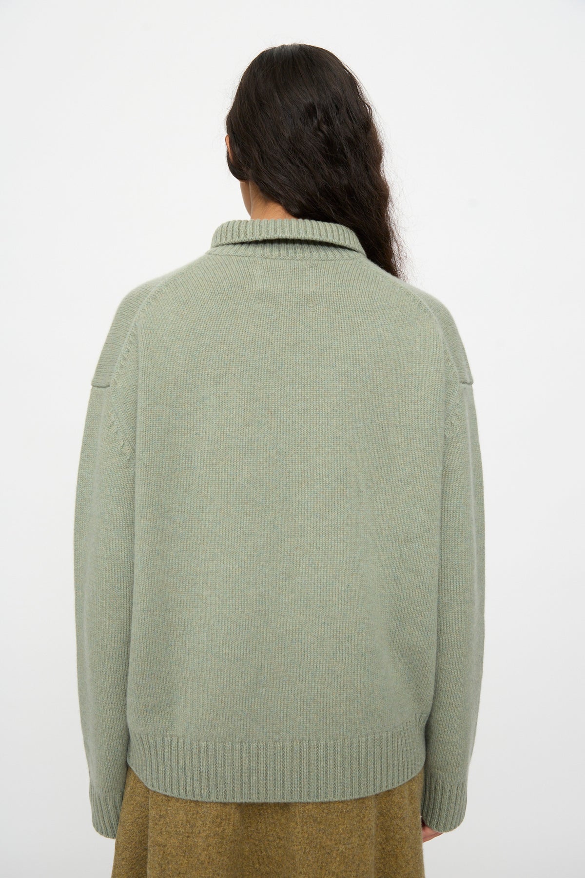 A person with long dark hair, standing with their back to the camera against a plain white background, is wearing the No. 514 Jan Sweater in Hemlock by Extreme Cashmere and an olive skirt.
