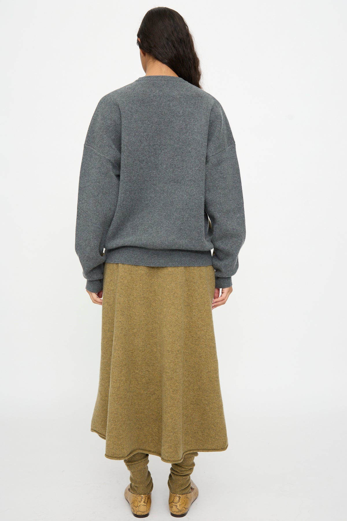 A person stands facing away against a plain white background, wearing the Extreme Cashmere No. 518 Trompe Sweater in Hunter and Felt—a loose knit gray sweater with a crew neckline—paired with an olive-green skirt, matching leggings, and tan shoes.