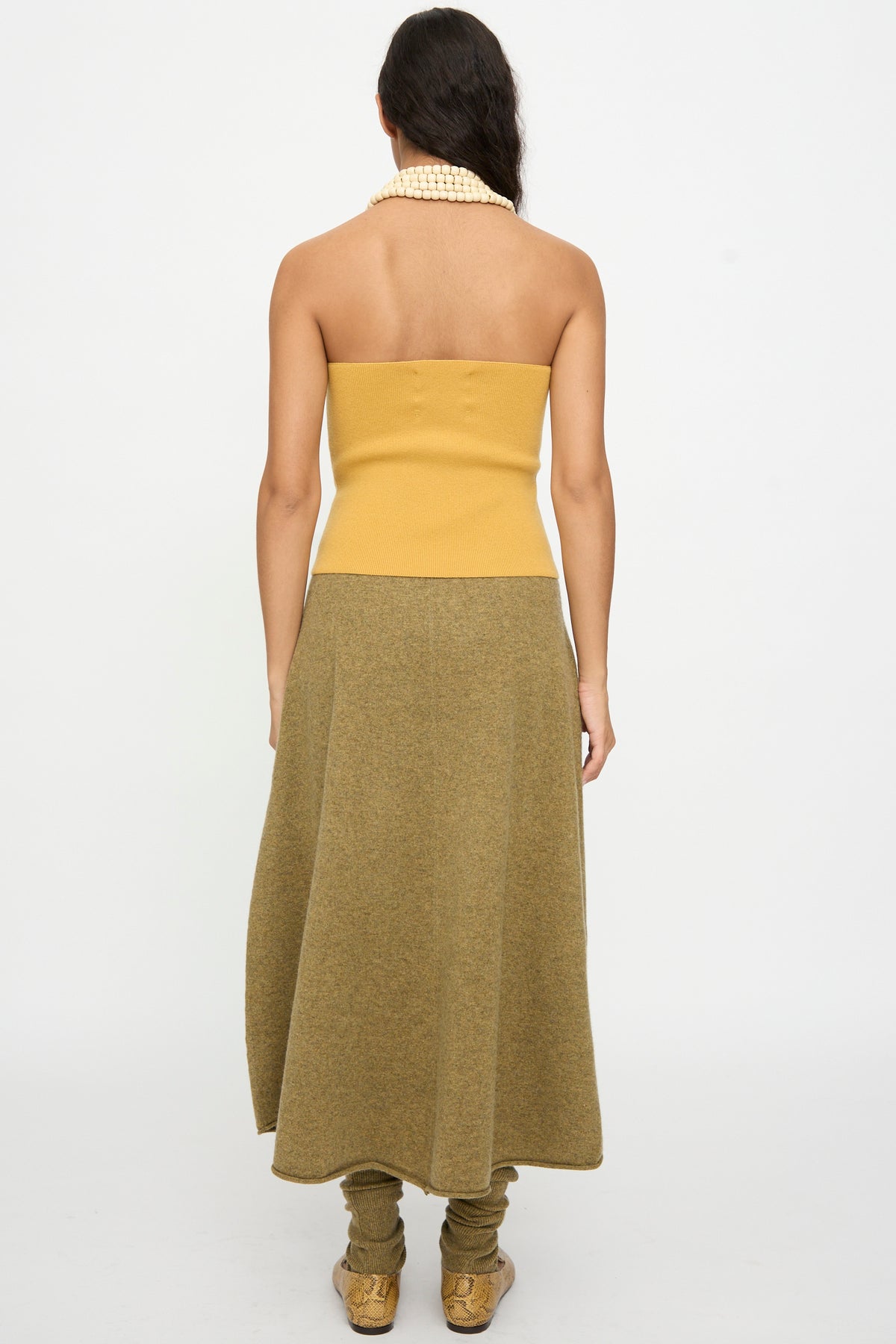 A woman stands facing away, wearing the Extreme Cashmere No. 519 Candy Top in Wheat, paired with an olive green midi skirt, pearl necklace, leg warmers, and sneakers against a plain white background.