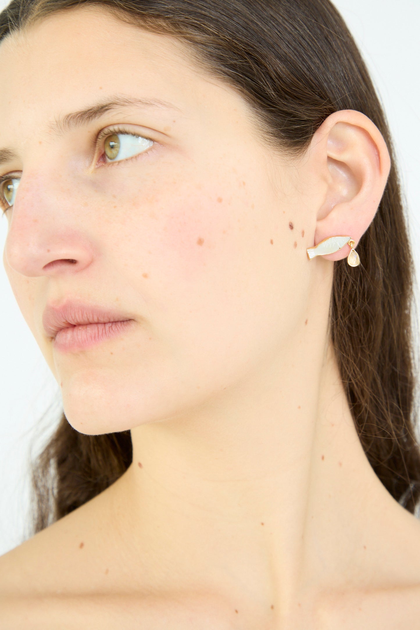 A person with long dark hair wears a left ear small opal Fish Drop Earring by Grainne Morton, with their gaze directed sideways.