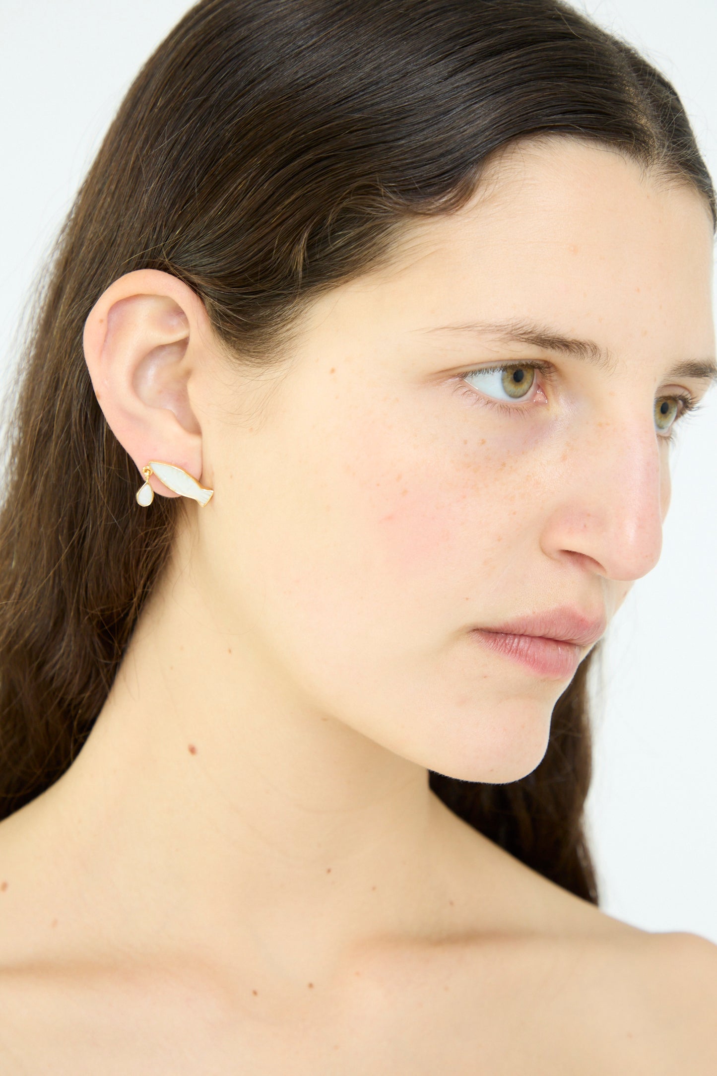 With long dark hair, the wearer showcases Grainne Morton's stunning Fish Drop Earrings in gold and white, adorned with delicate moonstone accents against a plain backdrop.