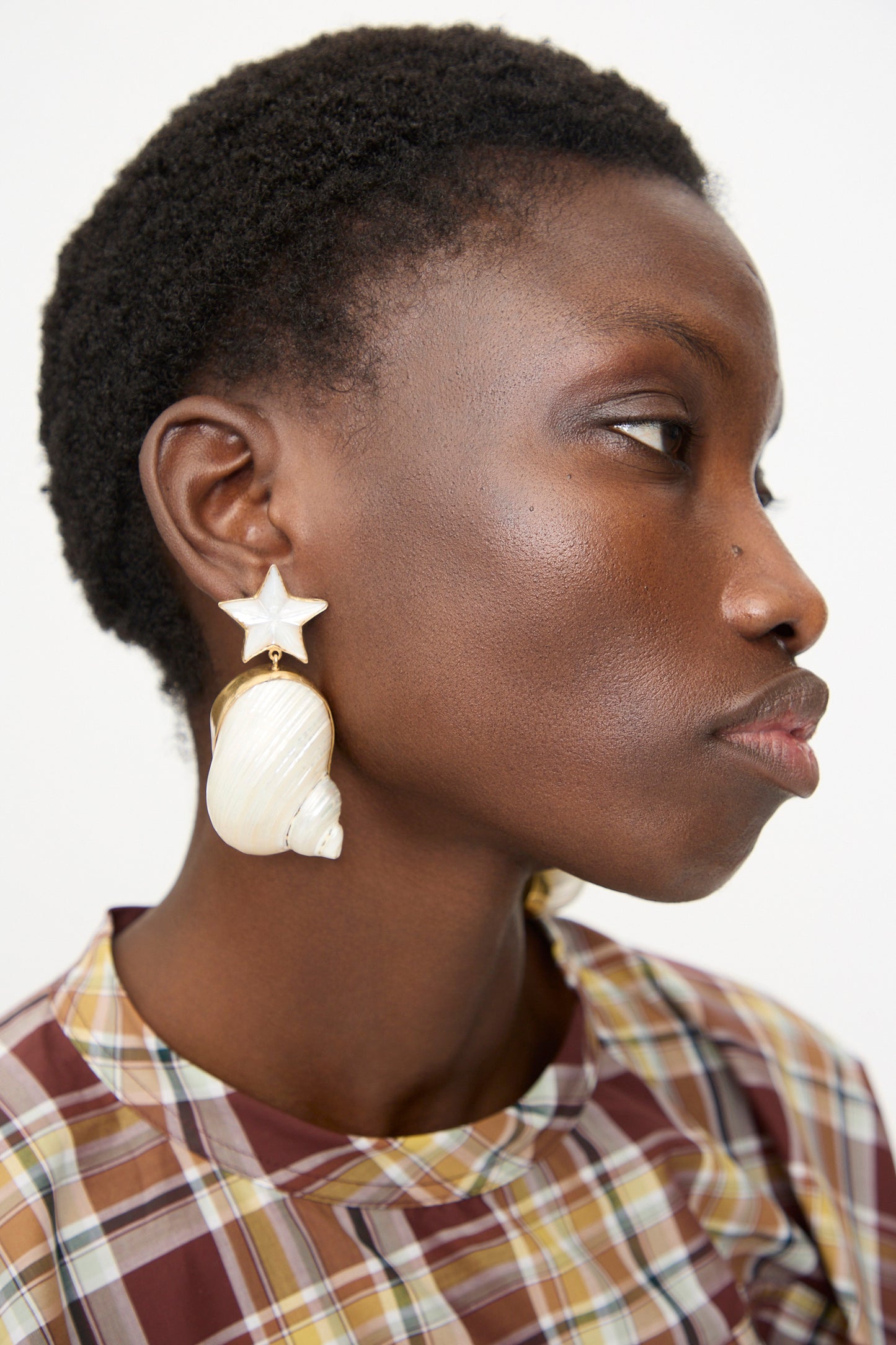 A person in a plaid shirt wears Grainne Morton Moon and Star Shell Drop Earrings—18K gold plated, seashell-shaped with a star accent—shown in profile against a plain background.