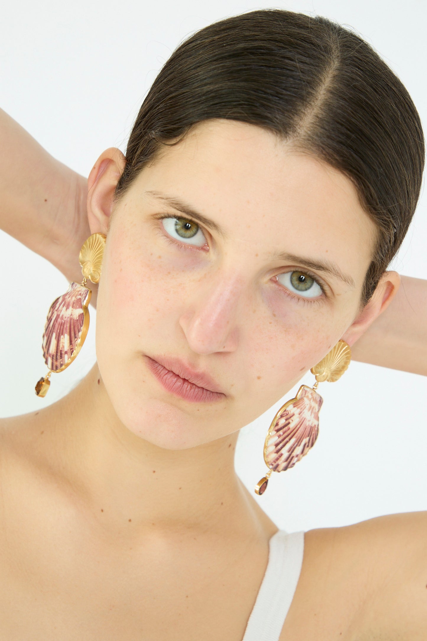 A person with dark hair poses with hands behind their head, wearing a white top and the elegant Grainne Morton Scallop Earrings inspired by ocean beauty, set against a plain background.