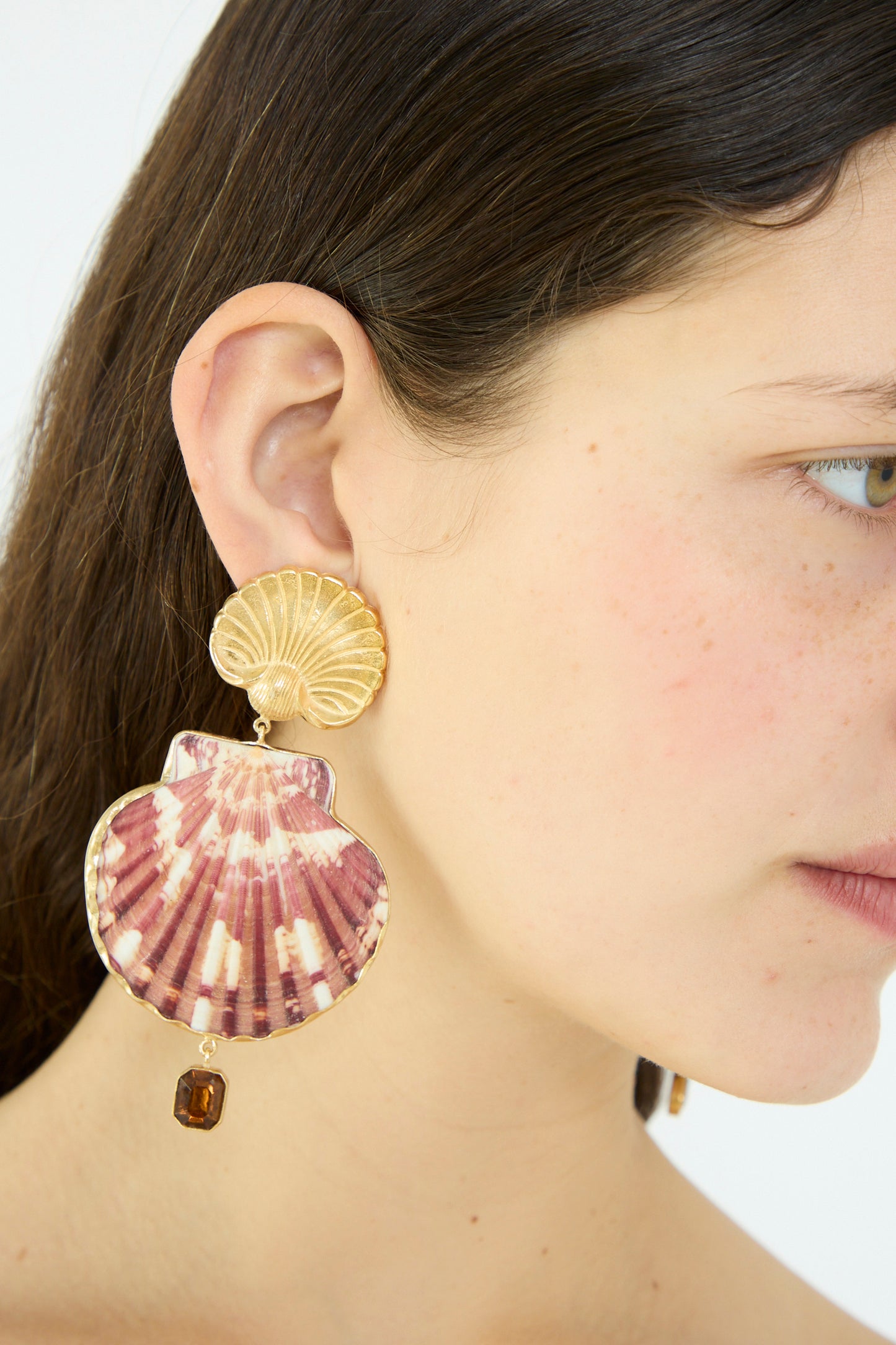 Someone wears a pair of Scallop Earrings by Grainne Morton, featuring large seashell-shaped drops with 18k gold-plated silver studs and brown gemstone details.