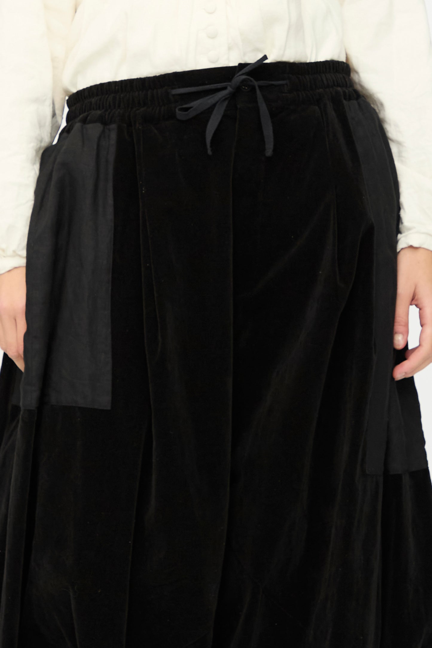 A person wears a white long-sleeve shirt and the Bloomer in Black skirt by Hallelujah, featuring velveteen cotton, an elastic waistband, and a drawstring bow at the center.