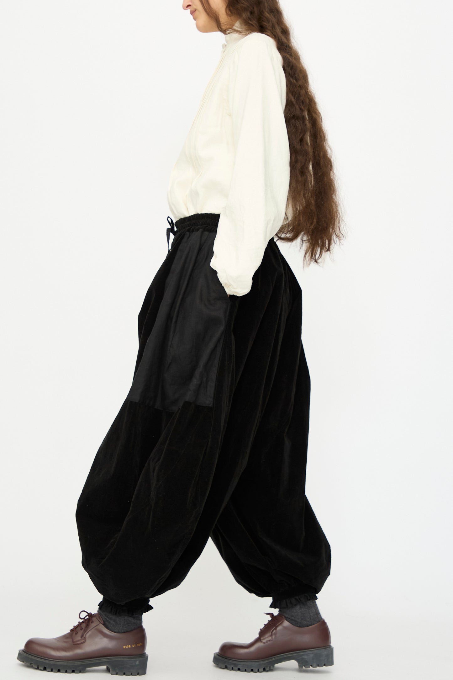 A person in profile wears the Hallelujah Bloomer in Black, a loose white shirt, dark socks, brown lace-up shoes, and has long brown hair, walking against a plain white background.