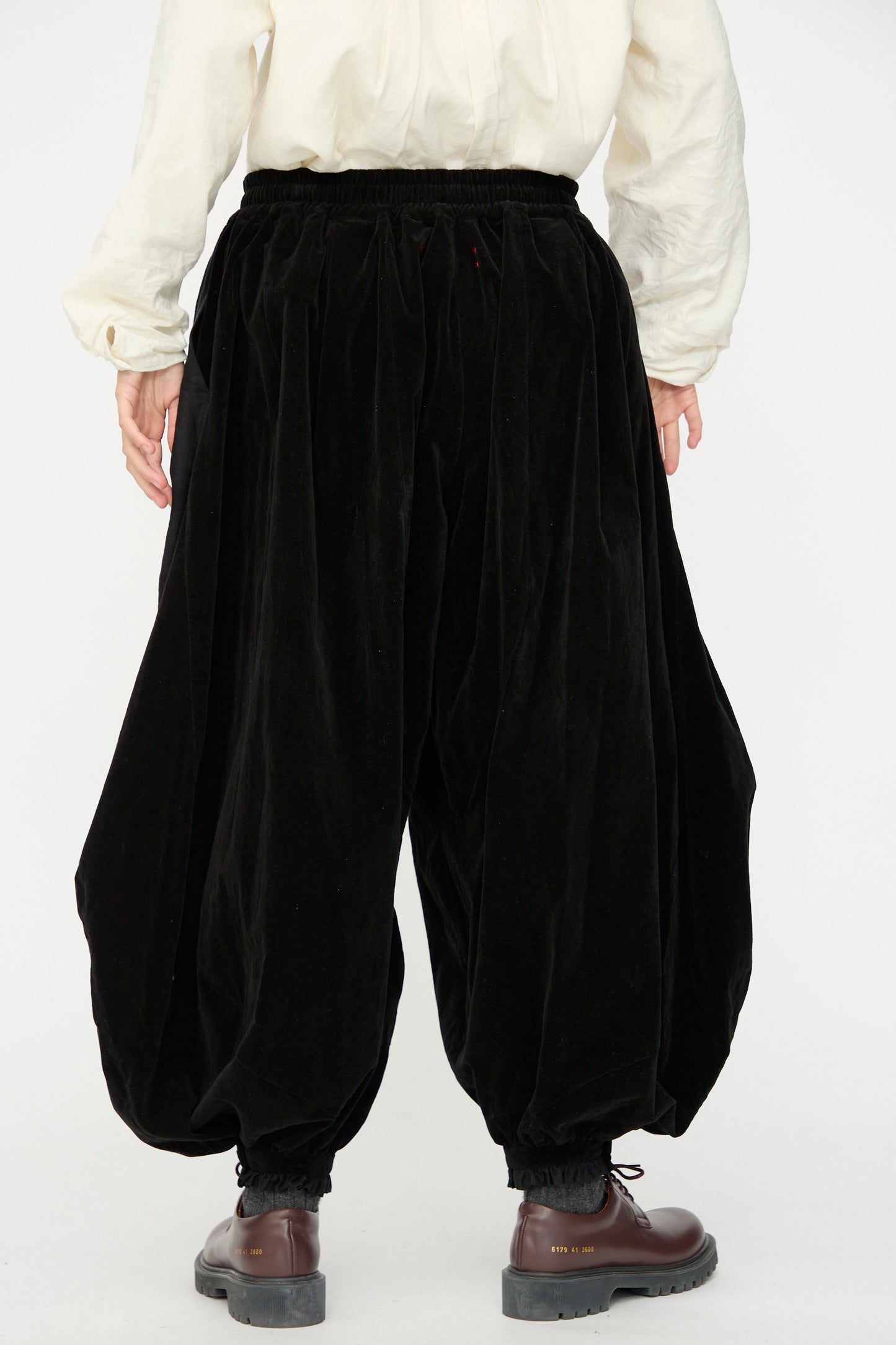 A person, shown from the back against a plain background, wears a white long-sleeve shirt, brown shoes, and Hallelujah’s Bloomer in Black—loose black velveteen cotton pants with balloon legs gathered at the ankles.