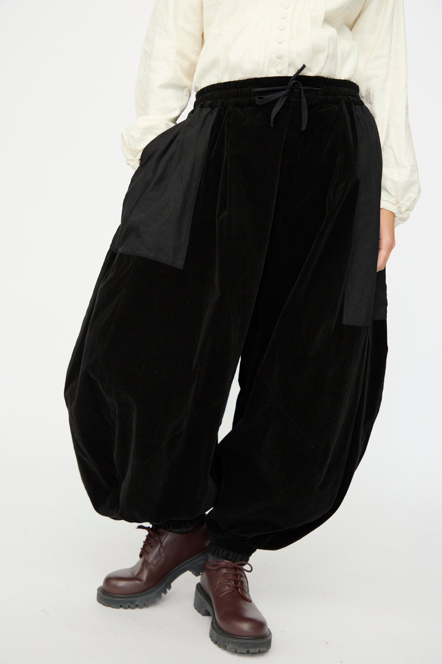 A person wearing the Hallelujah Bloomer in Black, featuring loose velveteen cotton and large front pockets, paired with a cream long-sleeve shirt and brown lace-up boots.