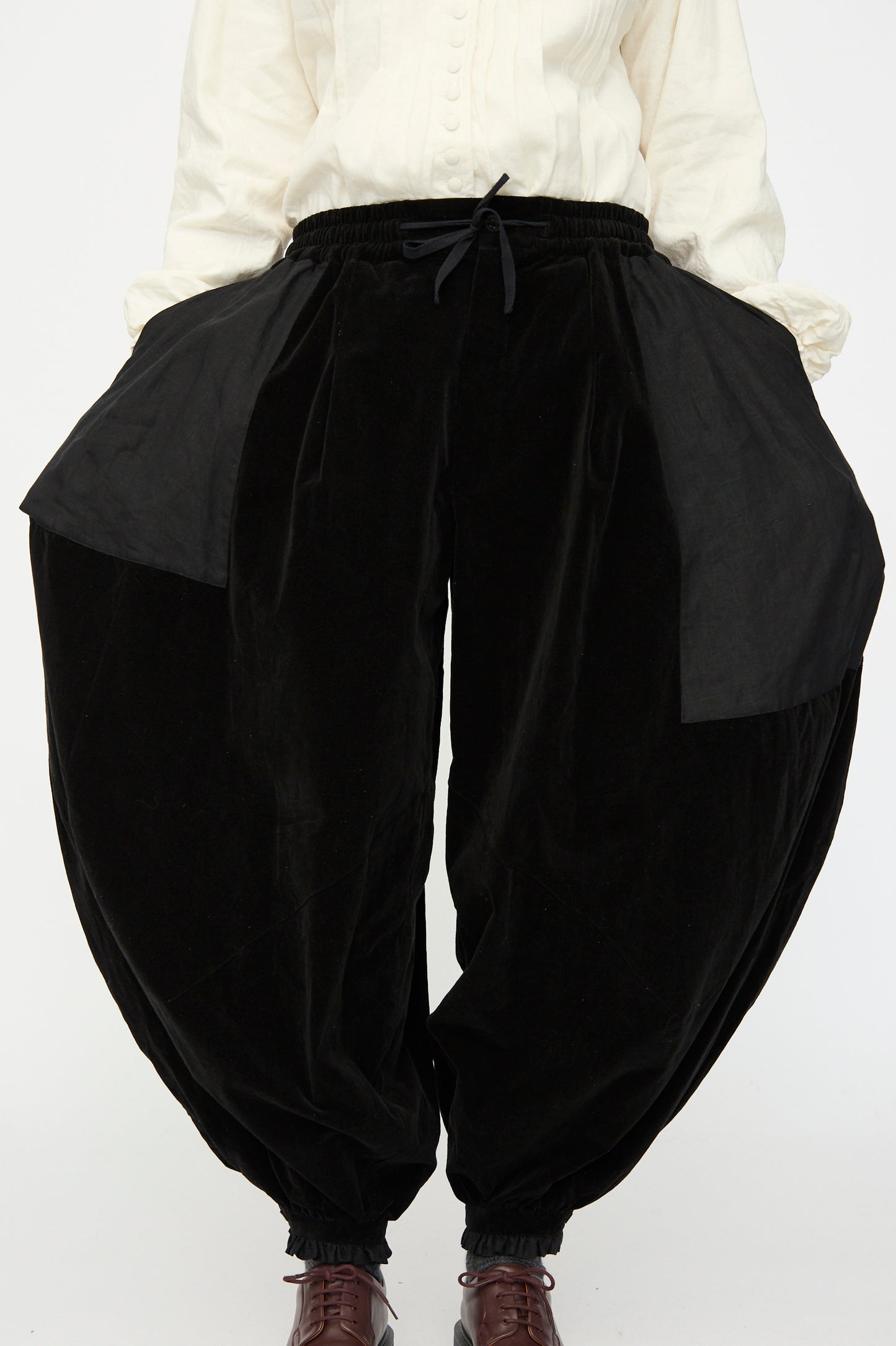 A person wears the Hallelujah Bloomer in Black—velveteen cotton pants with wide balloon legs, elastic cuffs, large side panels—paired with a white long-sleeve shirt. Their hands are in the pockets.