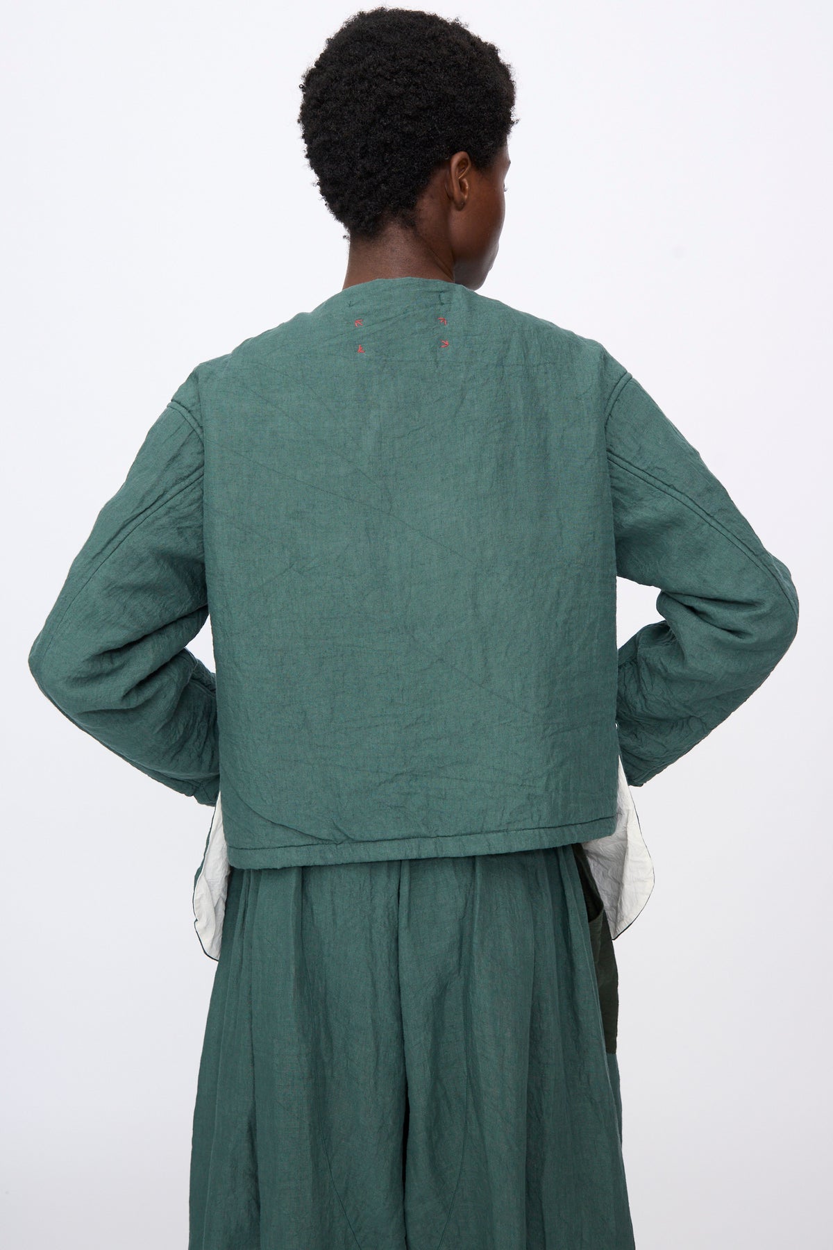 Back view of a person wearing a green, collarless, cropped quilted Hallelujah Linen Veste de Berger over a matching green skirt.