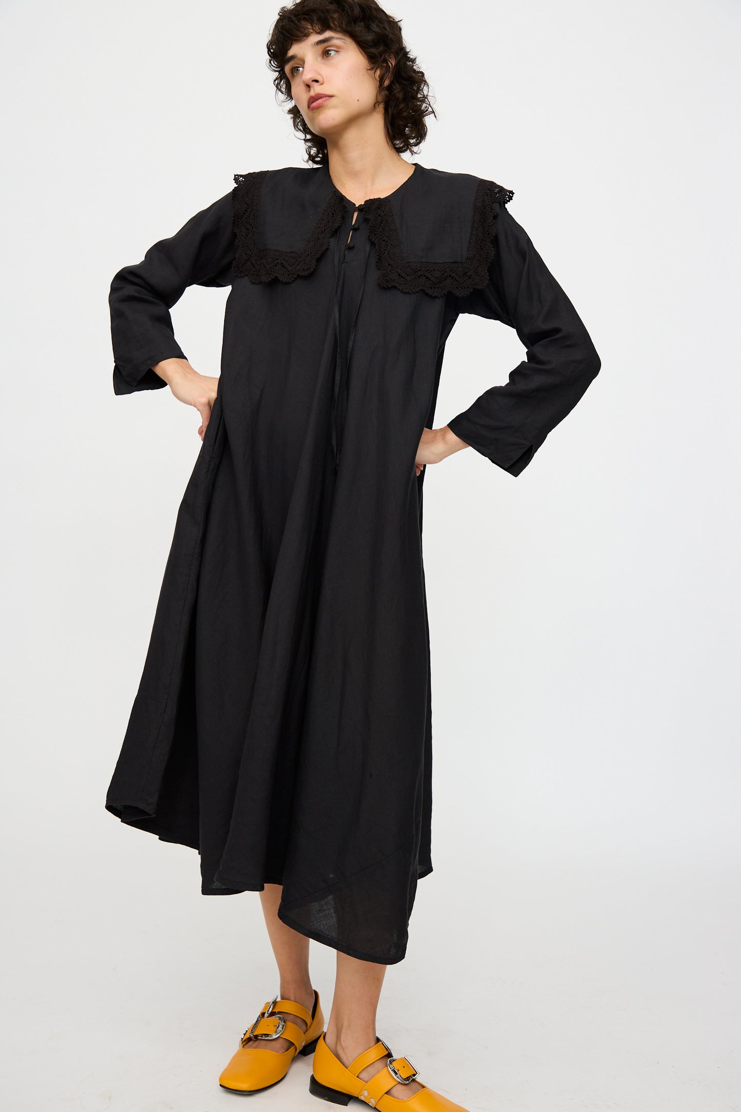 A person stands with hands on hips against a plain white backdrop, wearing the Robe Religieuse col carré in Black by Hallelujah. The black midi dress, featuring a large collar, is made from a linen/ramie blend and showcases an A-line silhouette, paired with yellow shoes.
