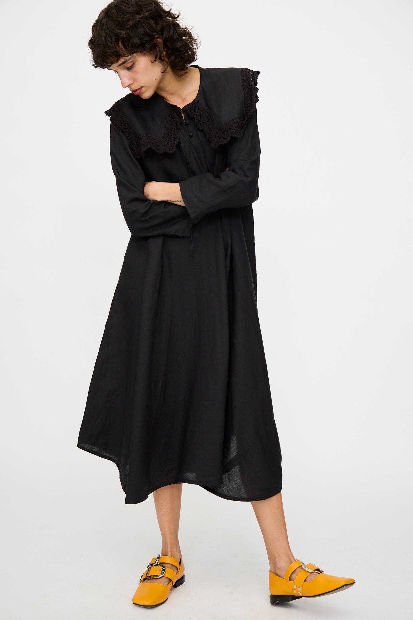 Someone wearing the Hallelujah Robe Religieuse col carré in black, paired with yellow pointed shoes, stands against a plain background.