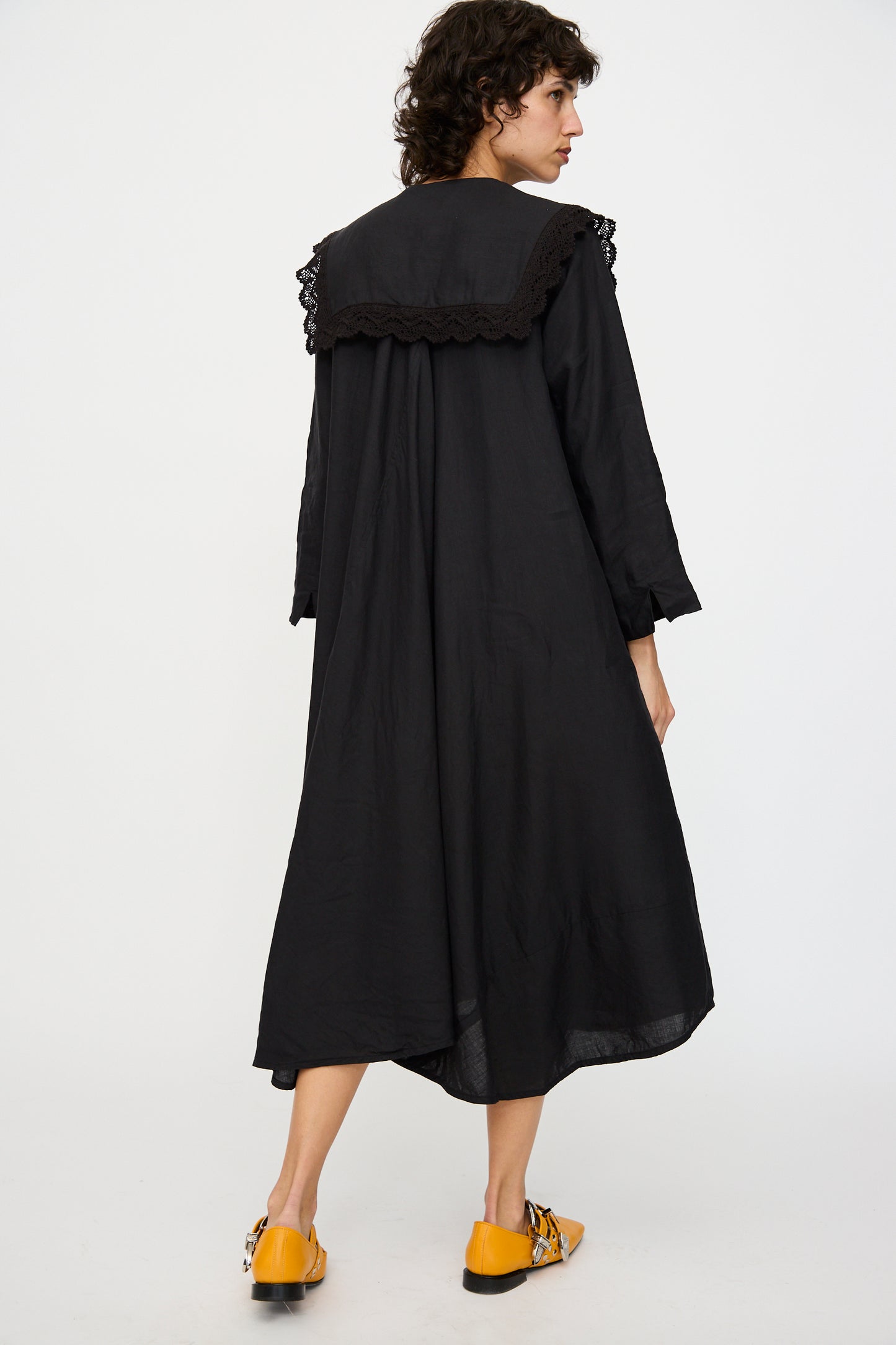 A person is seen from the back wearing Hallelujah's Robe Religieuse col carré in Black, a long midi dress with lace shoulder detailing, paired with vibrant yellow shoes against a plain white background.