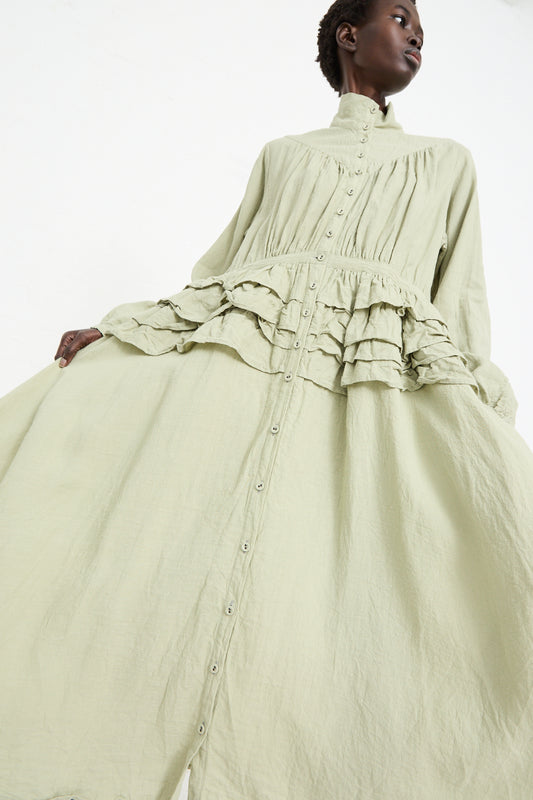 A person is wearing a Hallelujah Robe de Fille de Pionnier Dress in Sage with ruffled layers on the bodice and a high collar, showcasing an ancient Japanese dyeing method. They are looking upward.