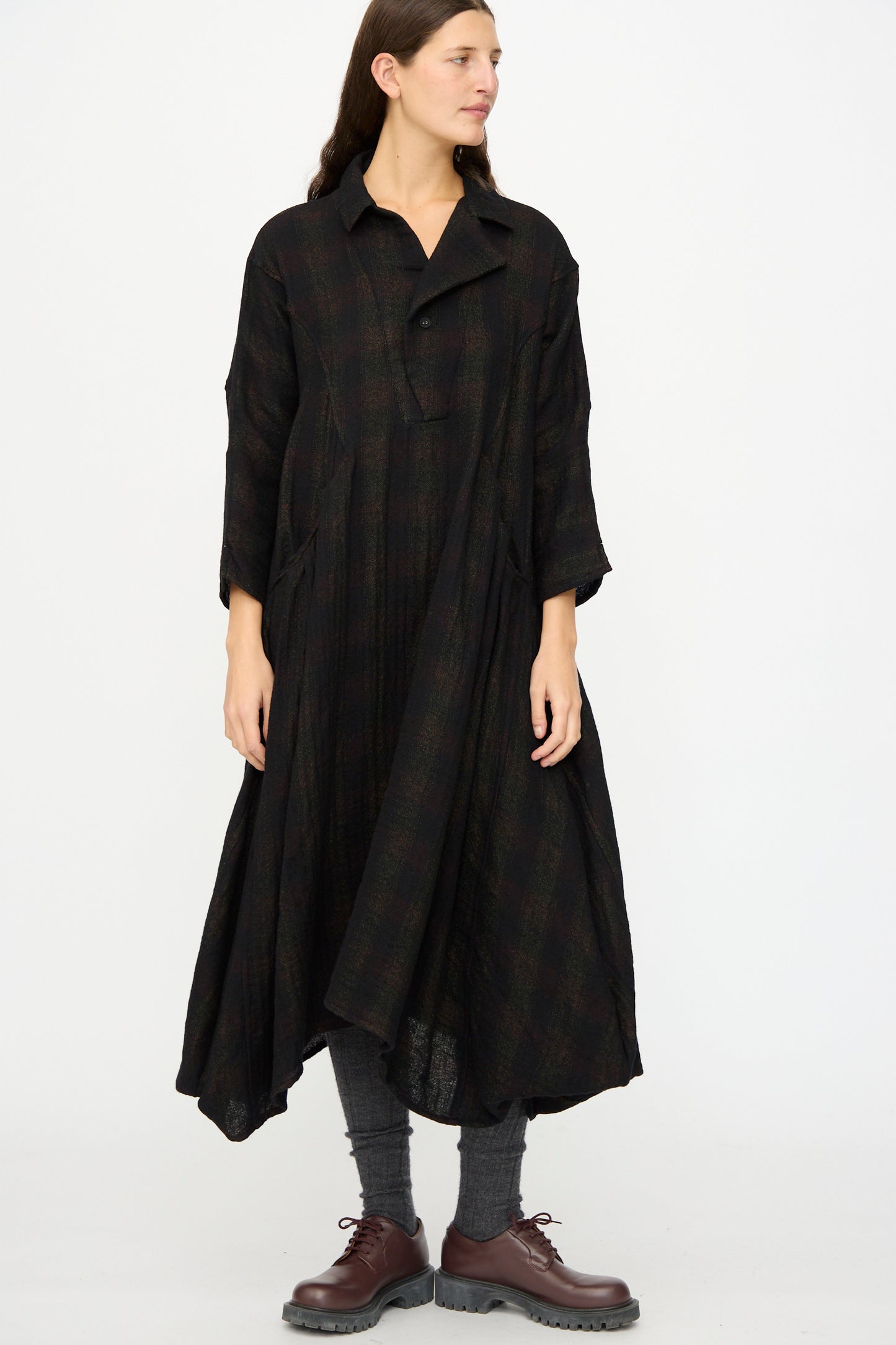A woman wears the Hallelujah Wool Cotton Robe de Berger in Black Mix, a loose-fitting, long sleeve checkered dress with a collar, styled with gray socks and brown lace-up shoes, made in Japan, set against a plain white background.