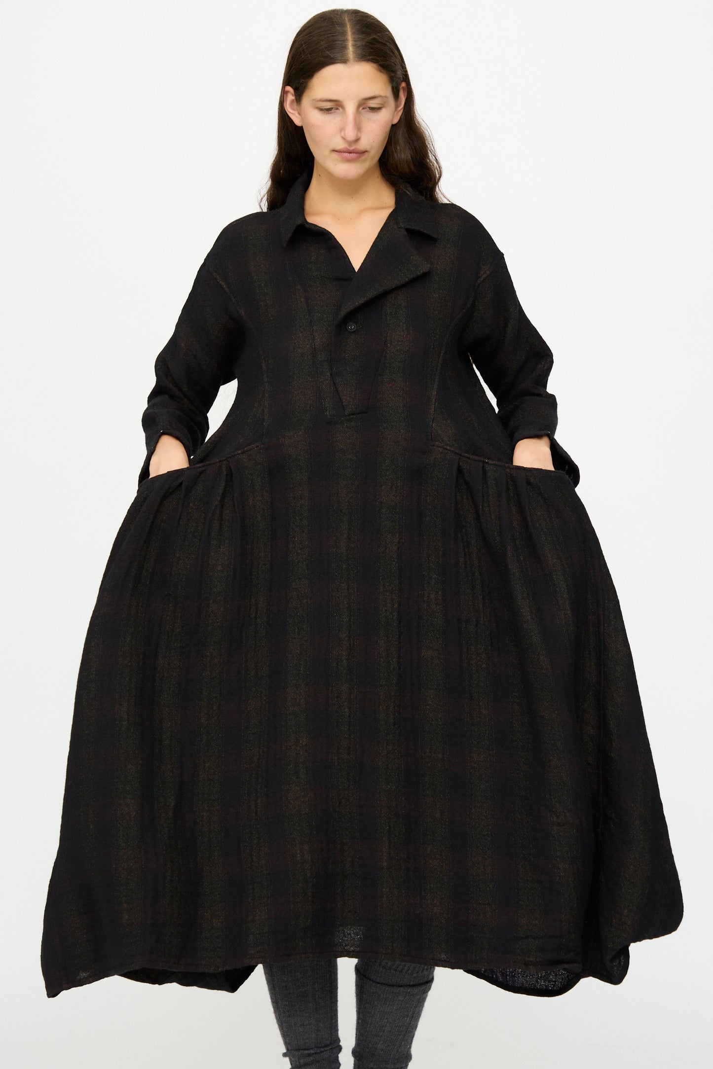 A woman stands against a white background wearing the Hallelujah Wool Cotton Robe de Berger in Black Mix, a loose-fitting, long-sleeve dress made in Japan with dark plaid, pockets, and paired with grey leggings.