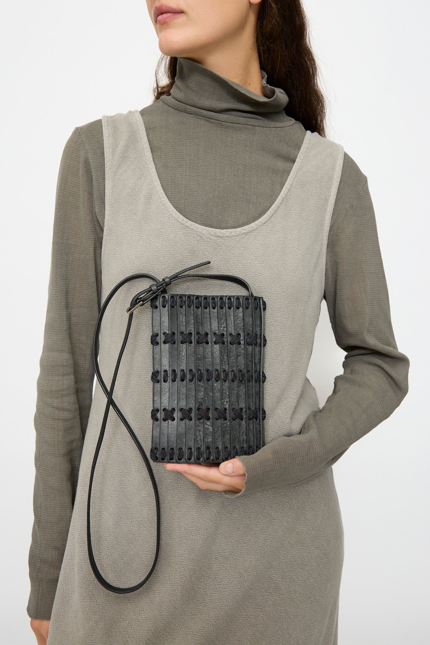 A woman in a grey turtleneck dress holds the Hatori Leather Phone 26 Bag in Waxed Coated Black with Indigo Lace, featuring a textured, woven design, handcrafted in Japan, against a plain background.