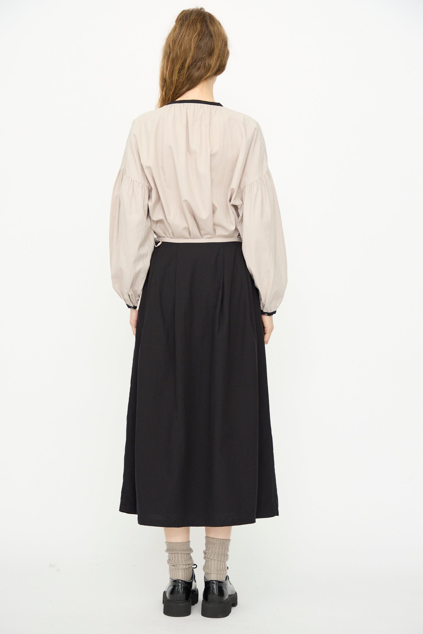 A person stands facing away against a white background, wearing a beige long-sleeve blouse, gray socks, black shoes, and the Ichi Antiquités Cotton Cashmere Viyella Skirt in Black.
