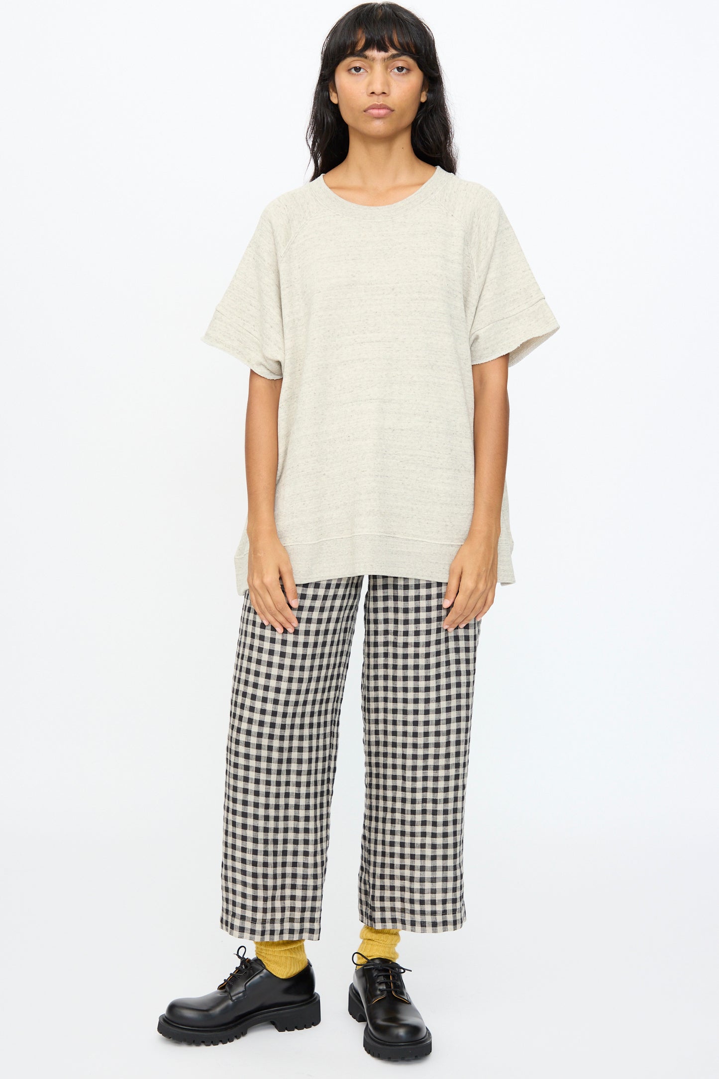 Person stands against a plain background, wearing the Ichi Antiquités Cotton Fleece Pullover in Gray, paired with black-and-white checkered pants, yellow socks, and black shoes.