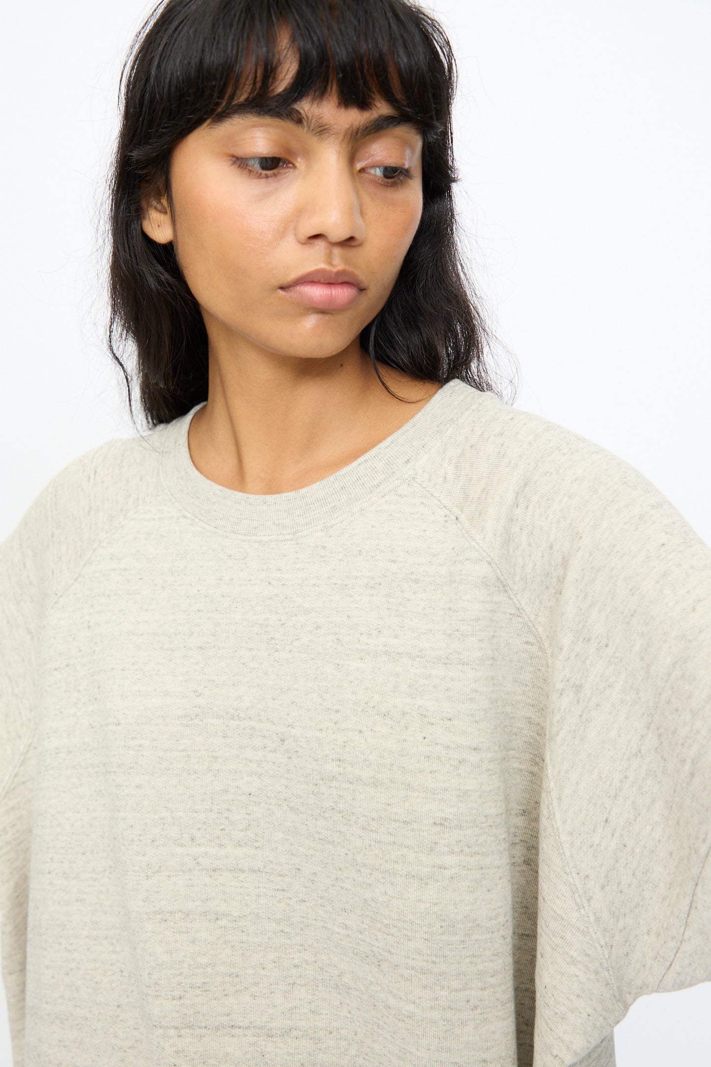 A woman with long dark hair and bangs wears the Ichi Antiquités Cotton Fleece Pullover in Gray, a loose-fitting style, as she looks slightly downward and to the side against a plain background.