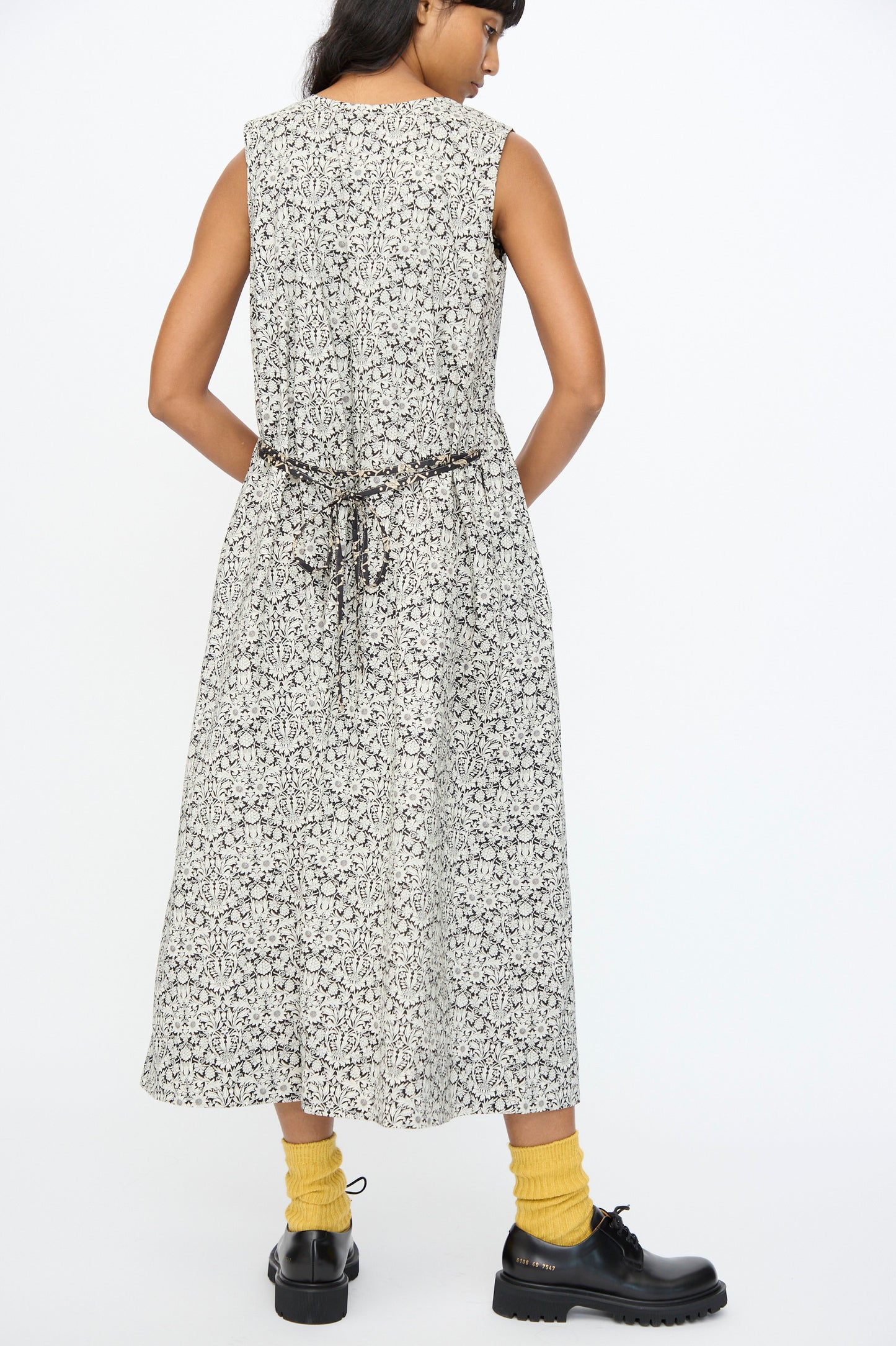 A person stands facing away, wearing the Ichi Antiquités Cotton Liberty Dress in Natural and Black, an ankle-length, sleeveless floral cotton dress with a thin belt, paired with yellow socks and black shoes.