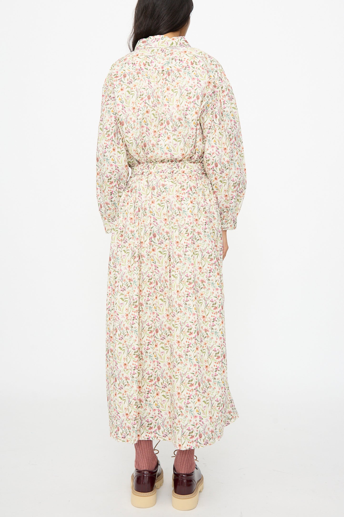 A person stands facing away against a plain white background, wearing a long-sleeved top, Ichi Antiquités Cotton Liberty Skirt in Flower Print with a belt, pink socks, and burgundy platform shoes.