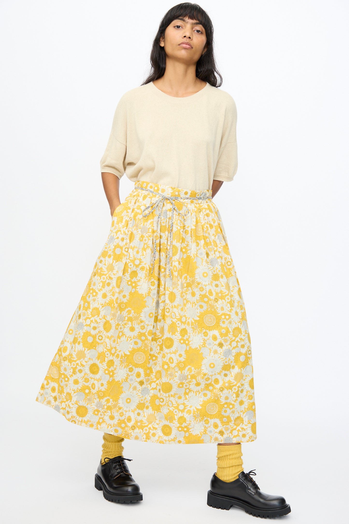 A person stands with hands in pockets, wearing a beige short-sleeve shirt, the Ichi Antiquités Cotton Liberty Skirt in Yellow, yellow socks, and black shoes against a plain white background.