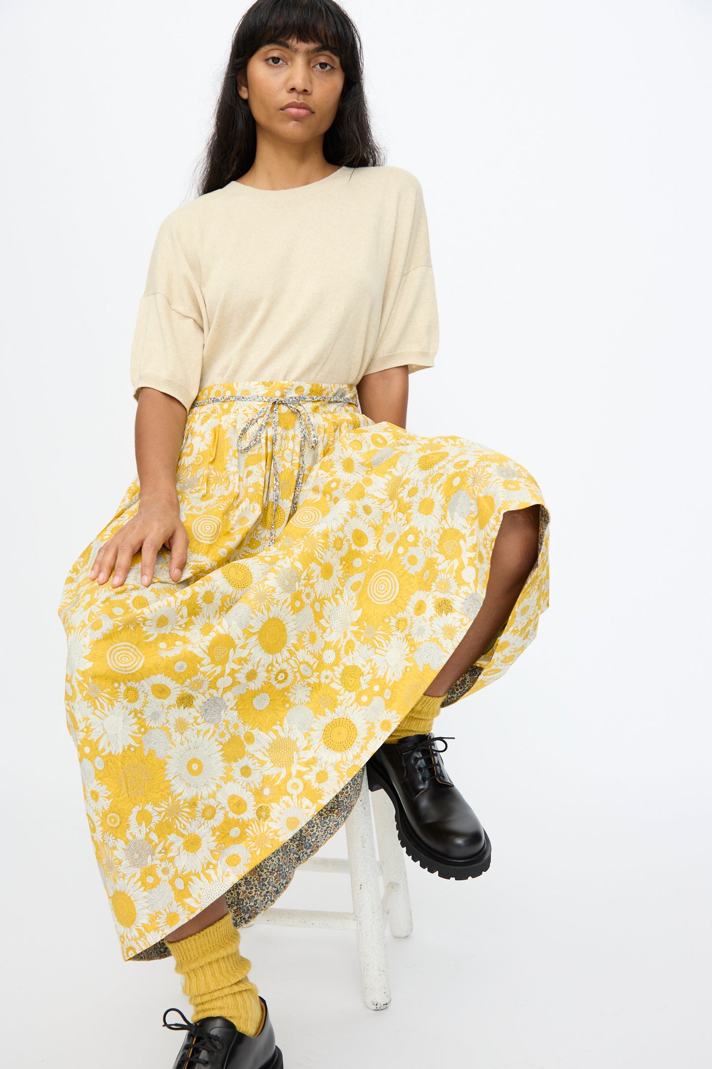 A person sits on a white stool wearing a beige t-shirt, the Ichi Antiquités Cotton Liberty Skirt in Yellow, yellow socks, and black lace-up shoes against a plain white background.