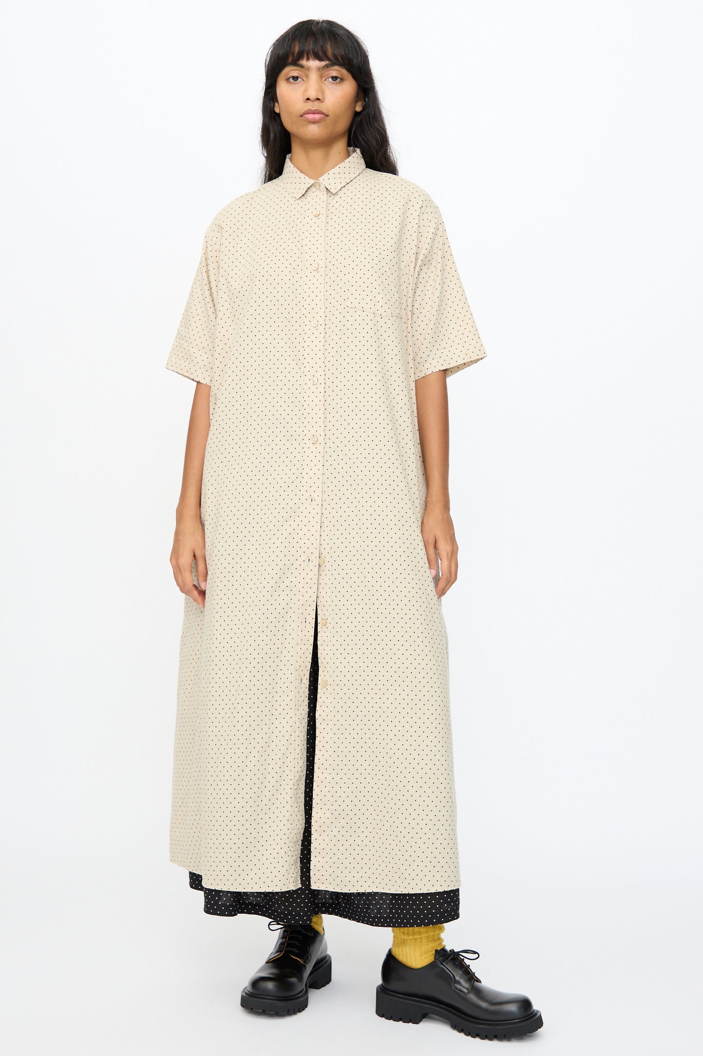 A woman stands facing forward in the Ichi Antiquités Cotton Linen Dot Dress in Natural—an oversized, beige, short-sleeve button-down dress with a subtle dot pattern—paired with black shoes, yellow socks, and a black layer underneath.