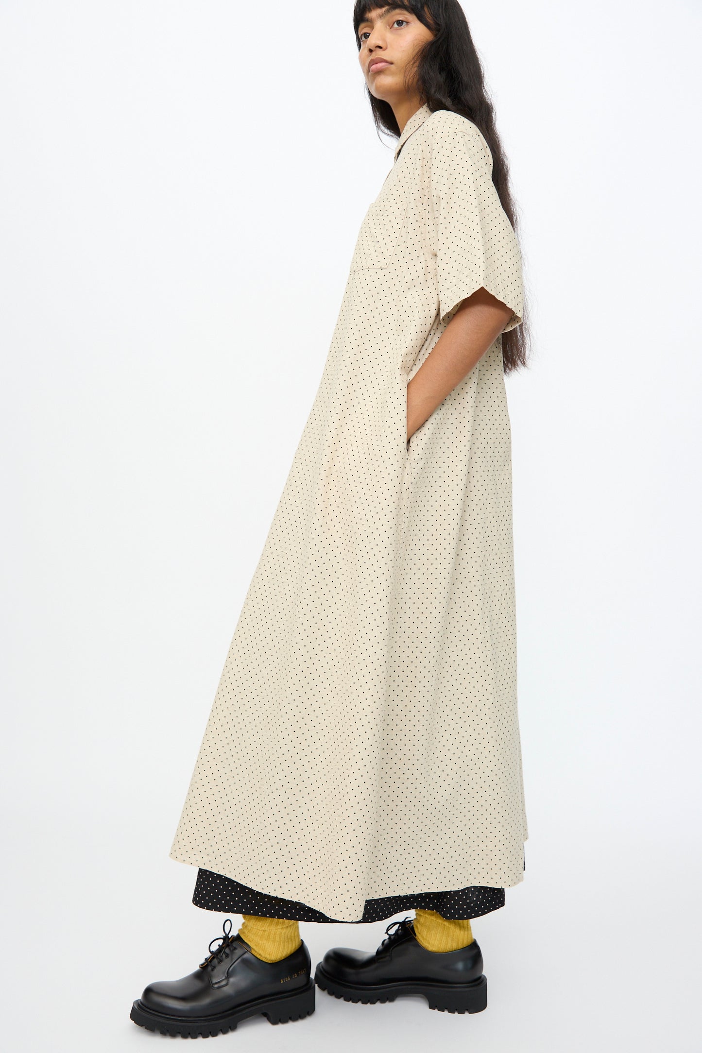 A person stands sideways wearing the Ichi Antiquités Cotton Linen Dot Dress in Natural, paired with black chunky shoes, yellow socks, and black pants peeking underneath.