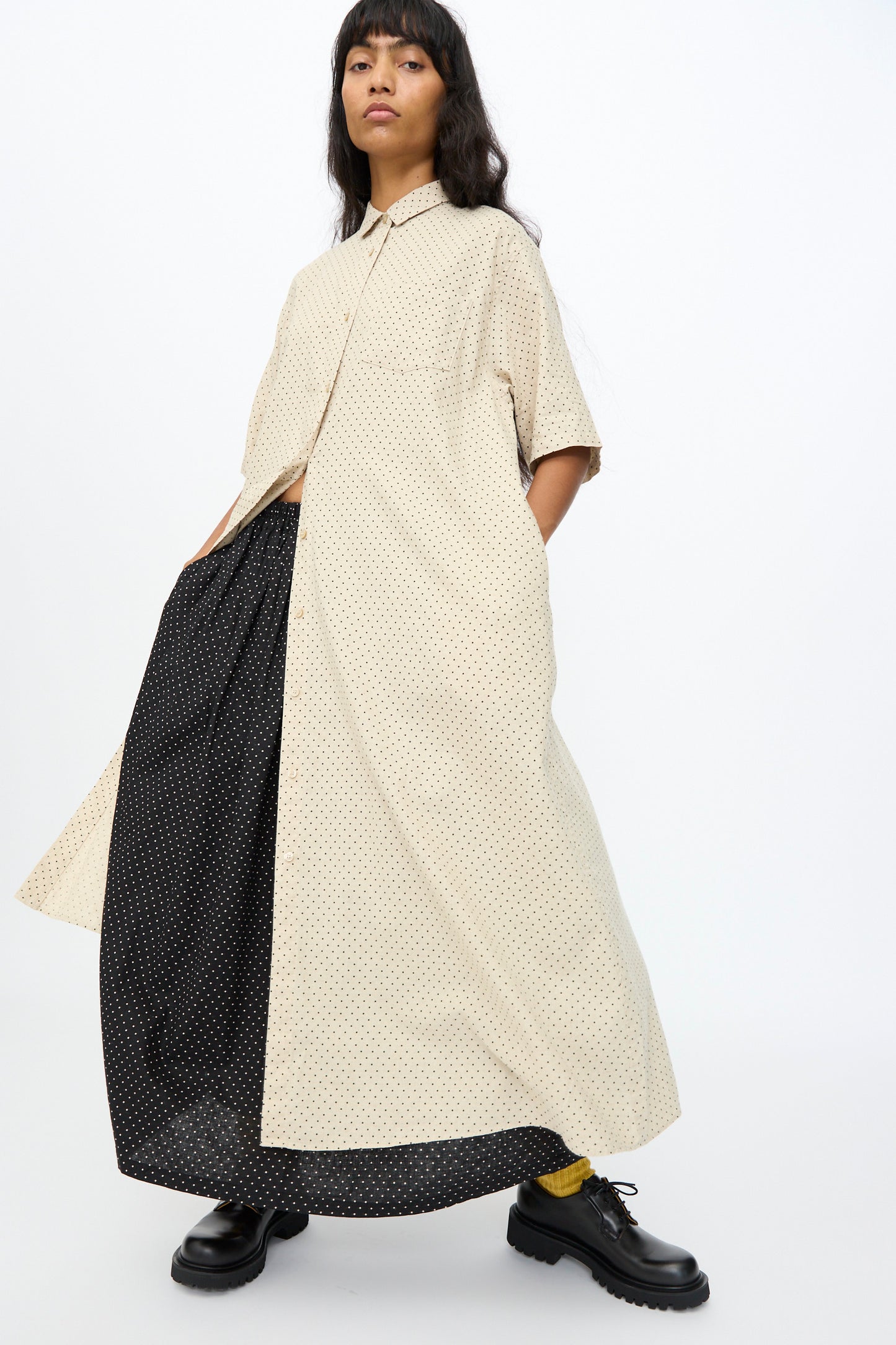A person wears the Ichi Antiquités Cotton Linen Dot Dress in Natural, oversized and short-sleeved, layered over a black polka dot dress with black shoes and yellow socks, standing against a plain background.